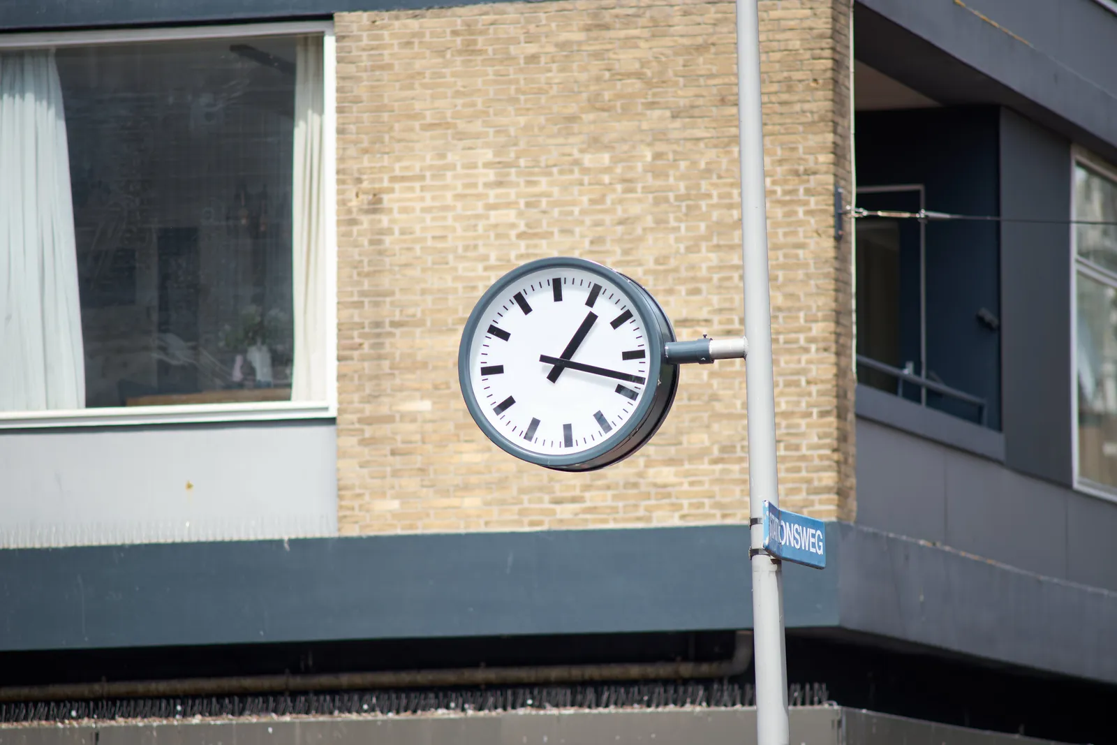 In the background there’s a brick building. In front of the building there’s a clock attached to a pole. It points at 18 past 1.