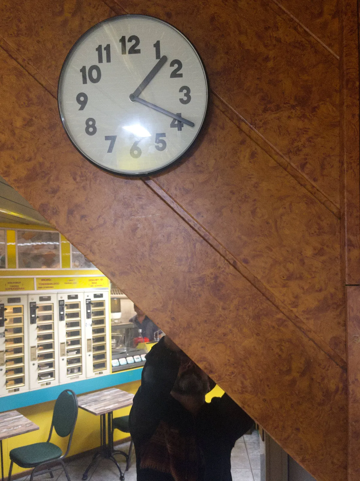 A picture taken in a Dutch snack bar. There’s a diagonal in the picture. The lower part of the diagonal is a mirror. In this mirror you can see one of those Dutch snack vending machines. The photographer is also visible. The upper part of the picture is part of a diagonal wall. There’s probably a staircase behind it. There’s a clock on this wall that points at 19 minutes past one.