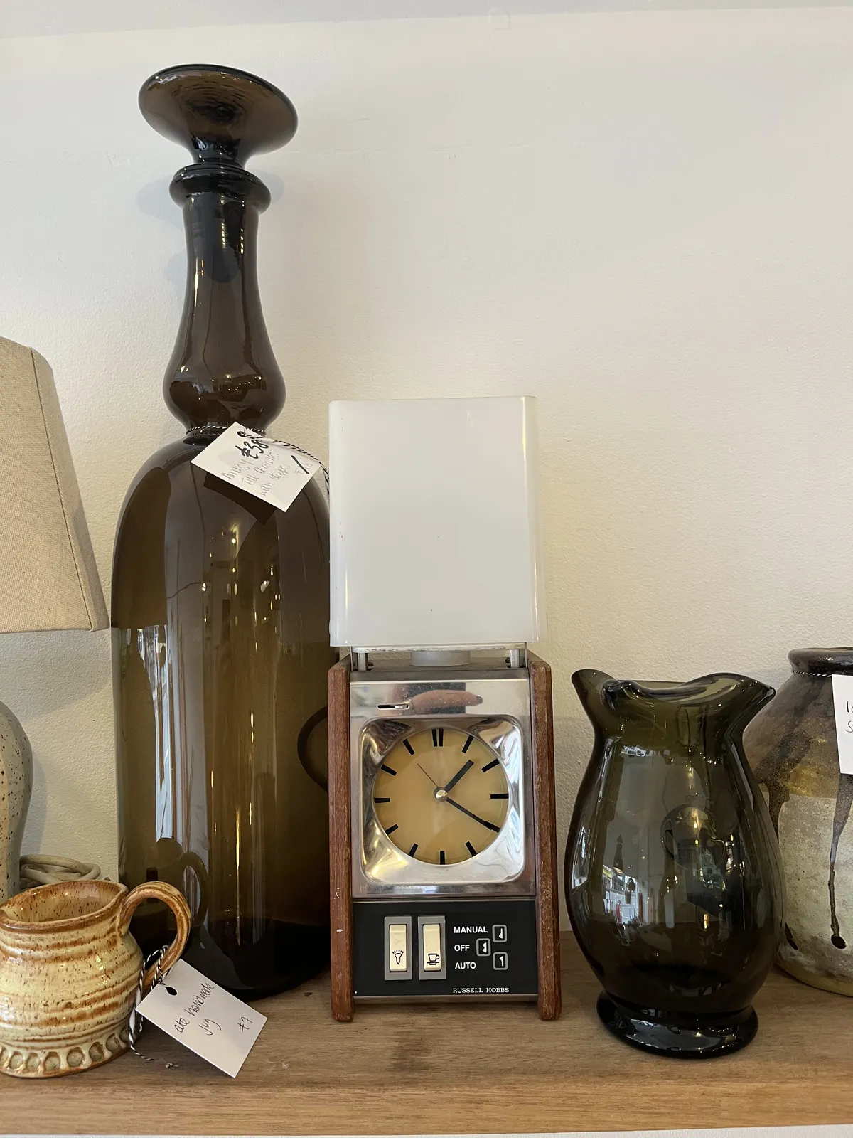 A few different vases, and a clock for sale in a thrift shop. The moniker Russel Hobs is on this clock. It has a few electrical switches on it. One of them is to control the light which is on top of the clock. The other one seems to be for switching on something like a coffee machine. The clock on this device points at 20 past one.  