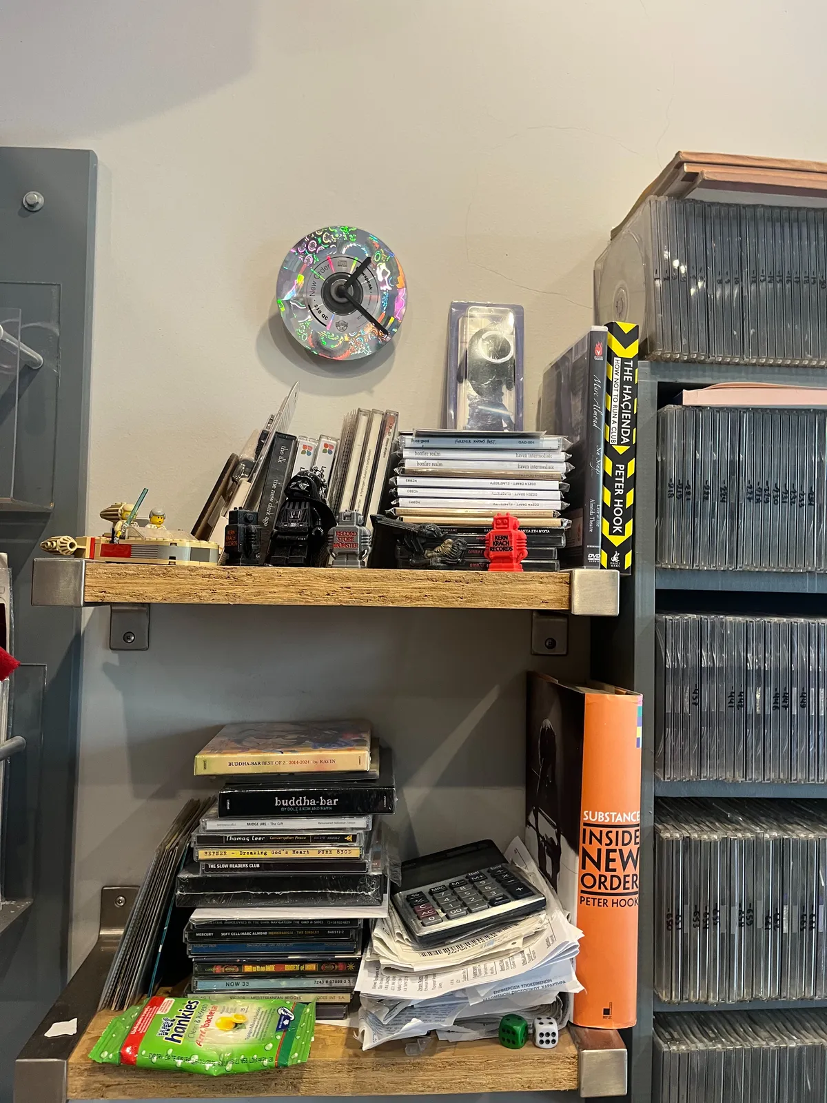 A clock made from a CD, pinned to a wall. Around it are CD boxes, some books, and a stack of special edition CDs. The clock points at 21 minutes past 1