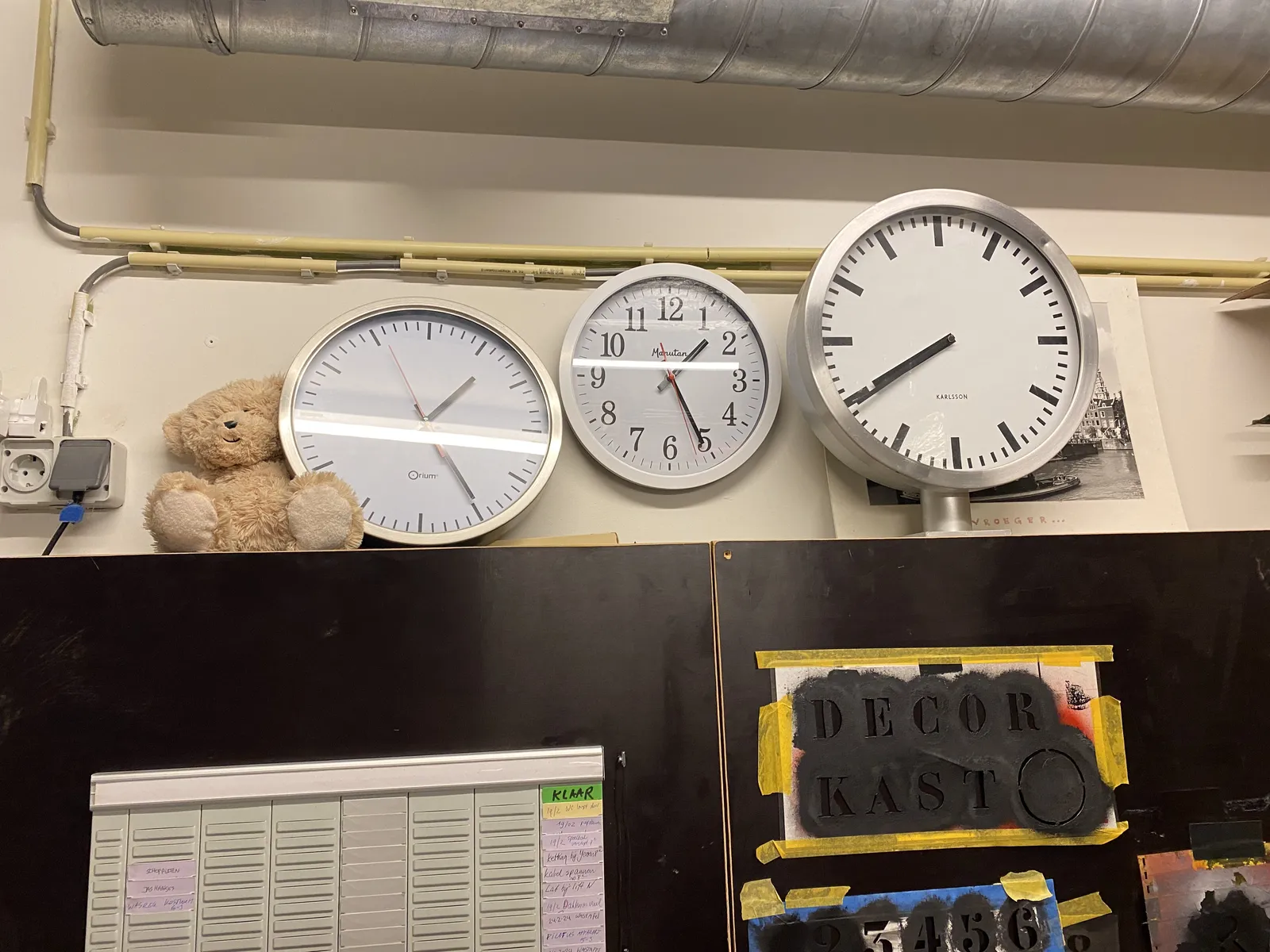 There are three clocks in this picture of a wall from what seems to be a theater set workshop. The clock on the left points at 24 minutes past one. The clock in the middle at 25 minutes past one. And then there's a clock on the right. Its hands are a bit off, so it could either point at 21 minutes to 8, or to 21 minutes to 9.