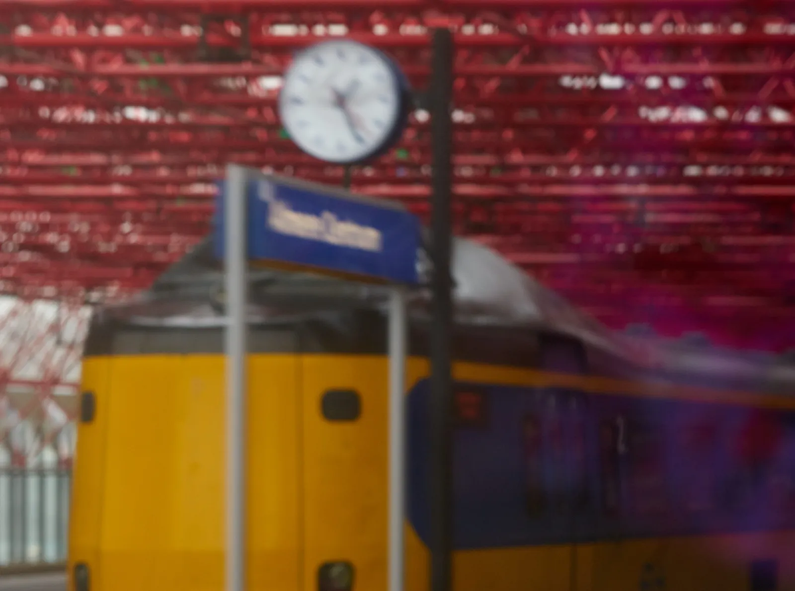 A blurry picture of a train in a station in The Netherlands, with a clock that points at approximately 26 past one.