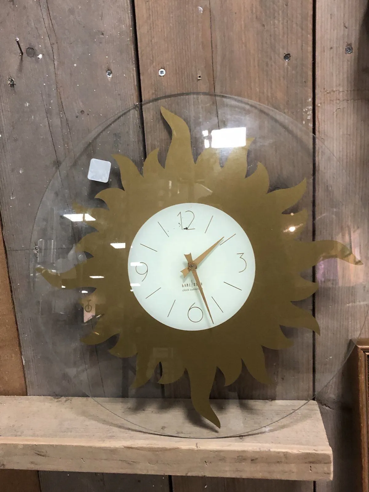 A clock made out of a round glass plate with a golden sun on it. It leans against a wooden wall. It points at 27 past 1.