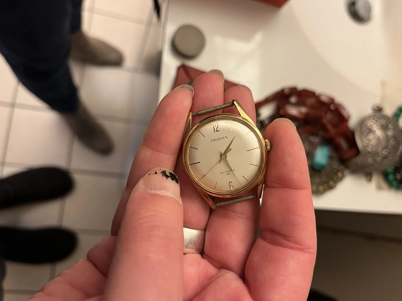 Somebody is holding an old wrist watch, without the bands, in their hand. In the background, looking down, are some blurry feet, more people seem to be watching this watch. The watch points at 27 minutes past one.