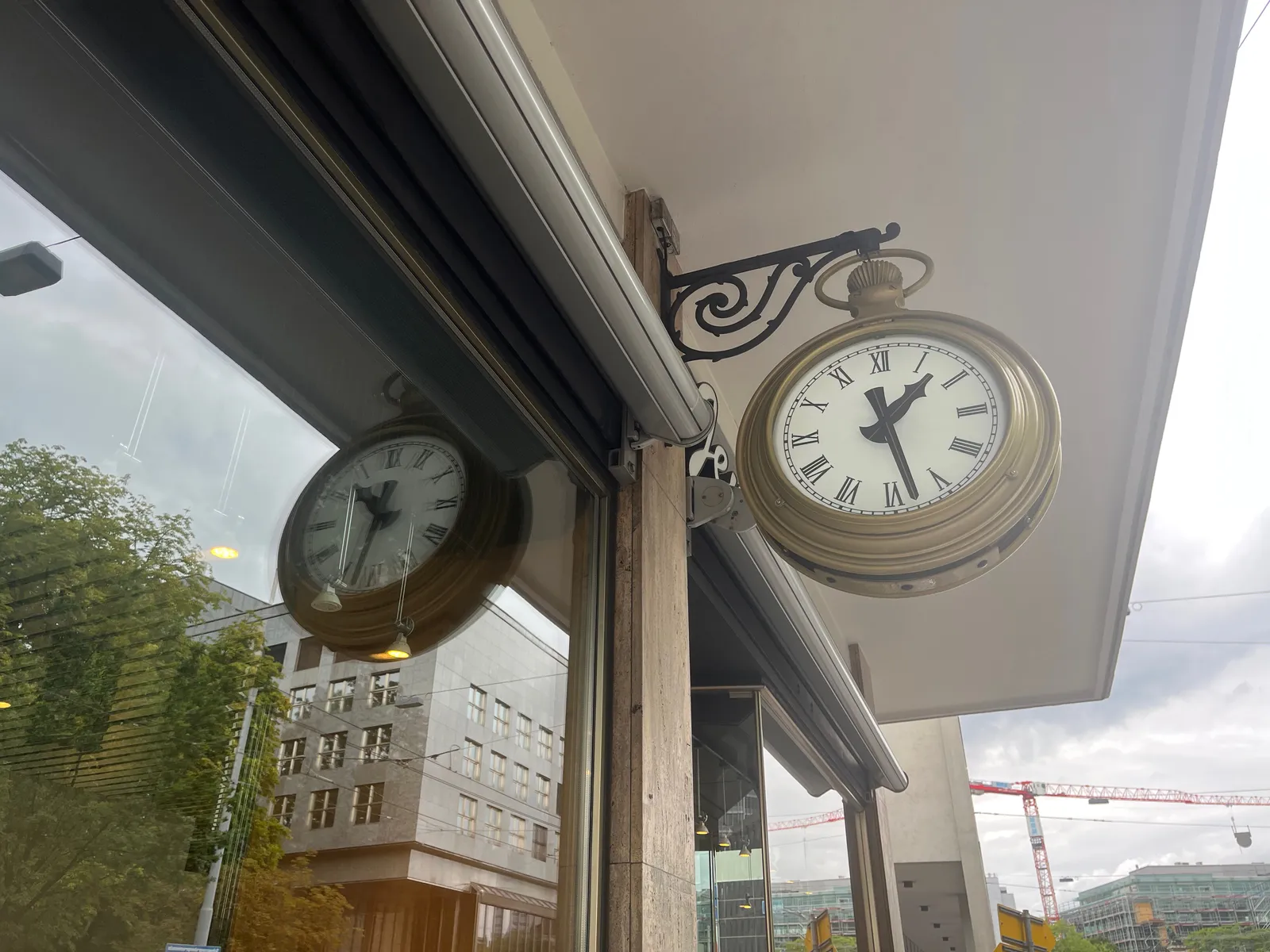 A clock attached to a building, and you can see its mirrored image in a shopping window. On the non-mirrored clock it’s 28 minutes past one. 