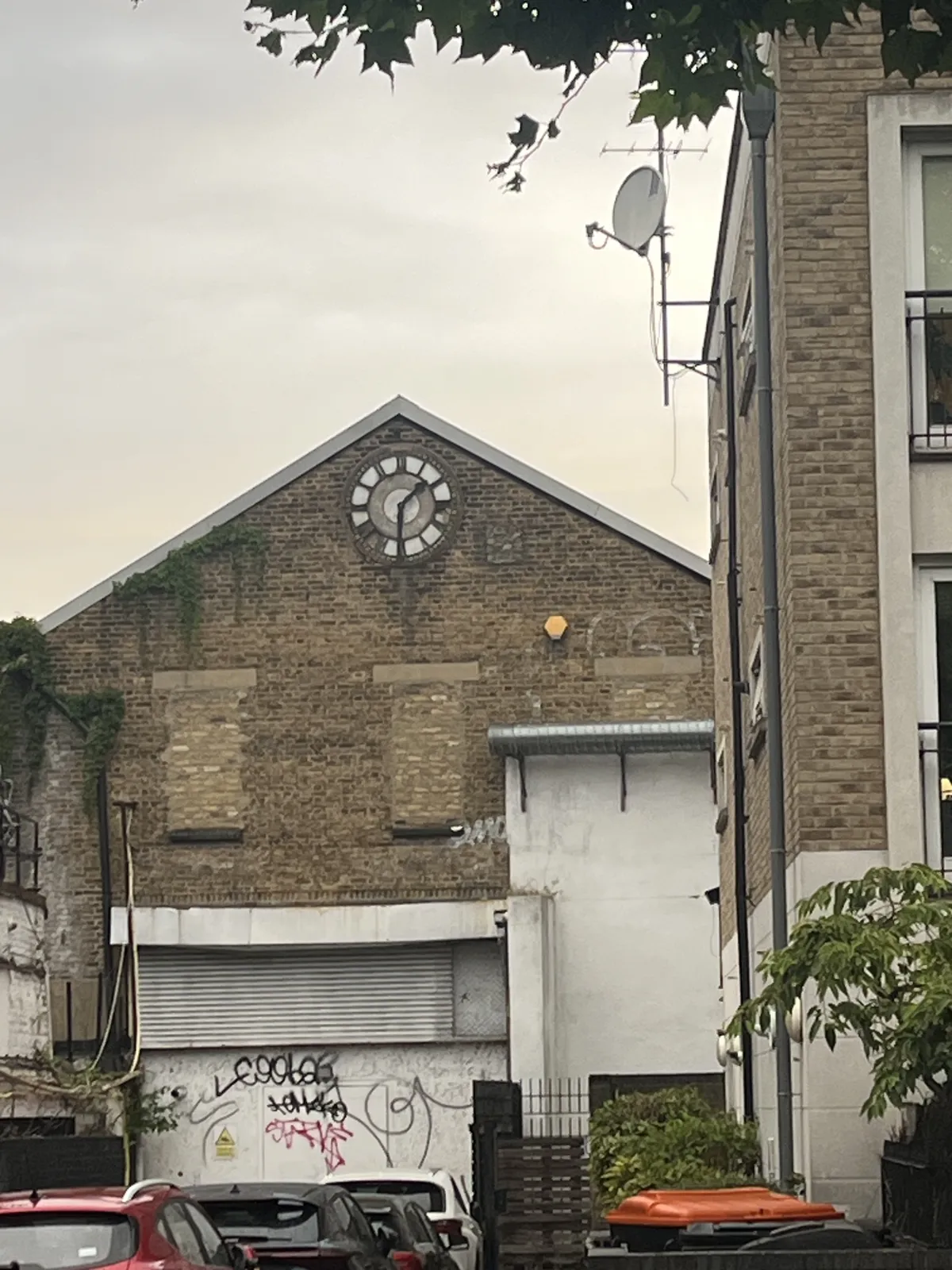 Four cars parked in front of a large industrial door on an old building. There’s a clock on to pof this building. It points at half past one.