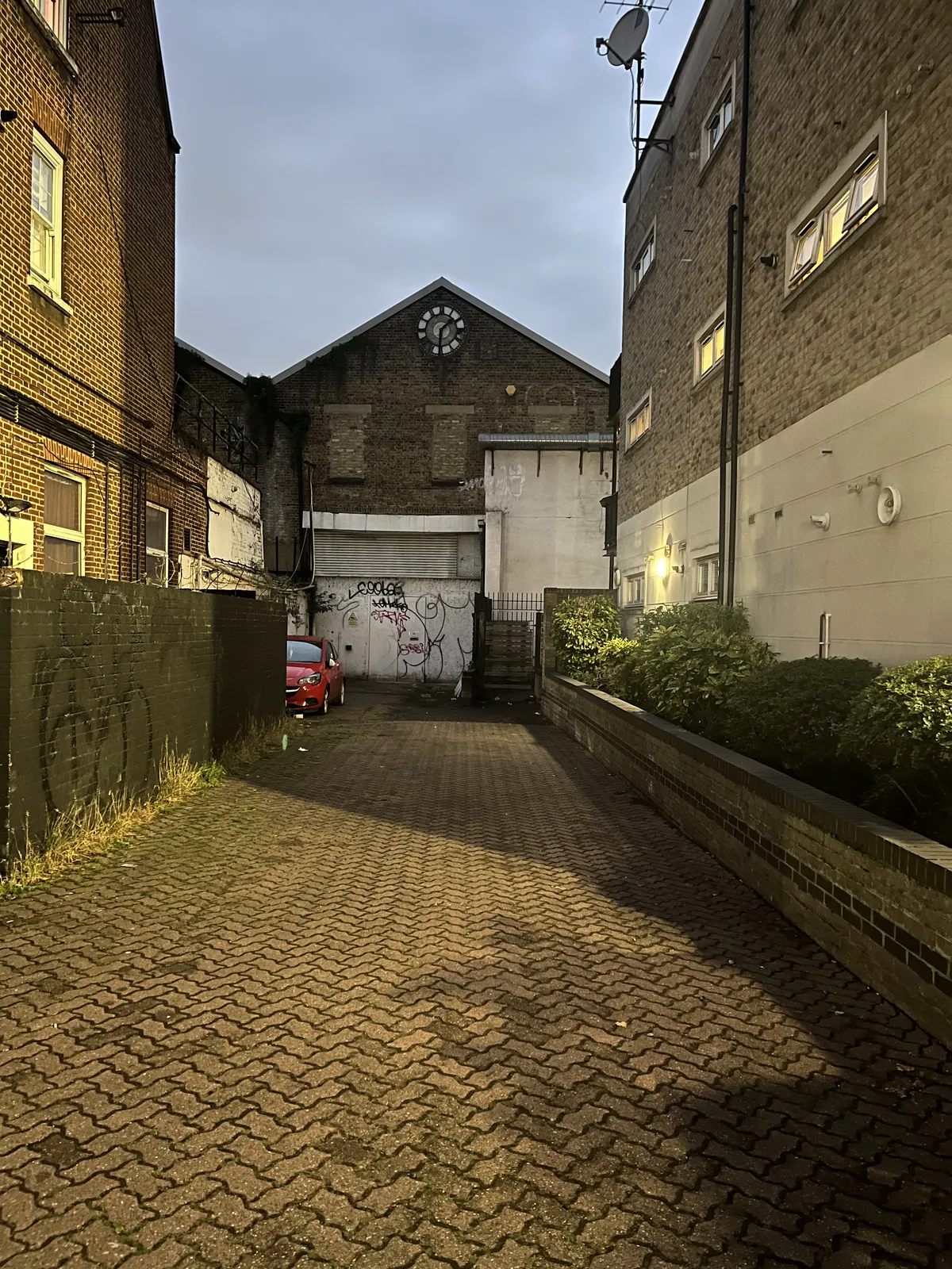 Looking into an alleyway, at an old brick industrial building. There’s a clock on this building which points at approximately 31 minutes past one.
