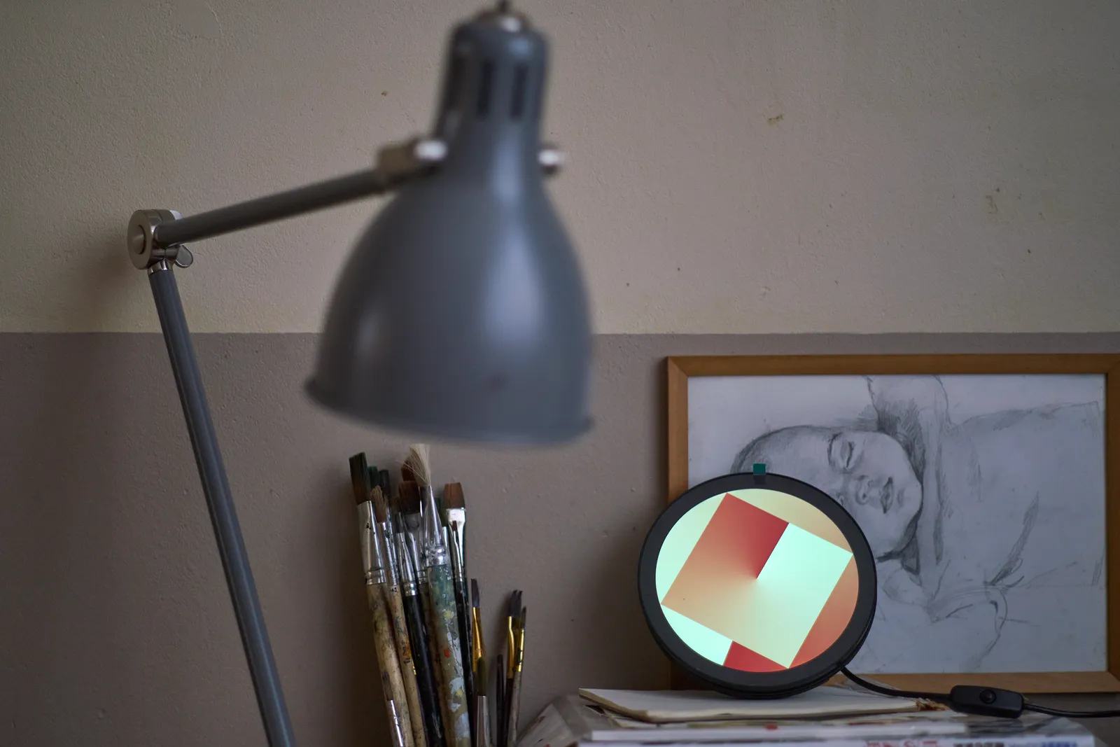 A classical lamp, out of focus in the foreground. In the background there are quite a few paint brushes, and a round display, in front of a drawing of a sleeping baby. On the display there’s a square with a gradient in it, and the same gradient is in the background. If you want to you might see a clock in these two gradients. It points at approximately thirty three minutes past one.