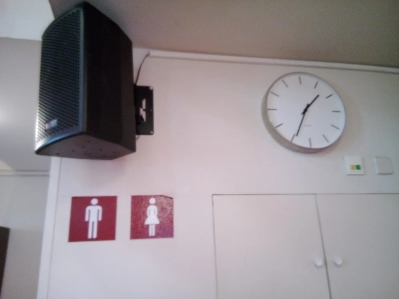 Below a speaker there are two stickers with the classic, minimalist icons of a man and a woman. Next to the speaker there’s a clock that points at 34 minutes past one. The picture is a bit blurry.