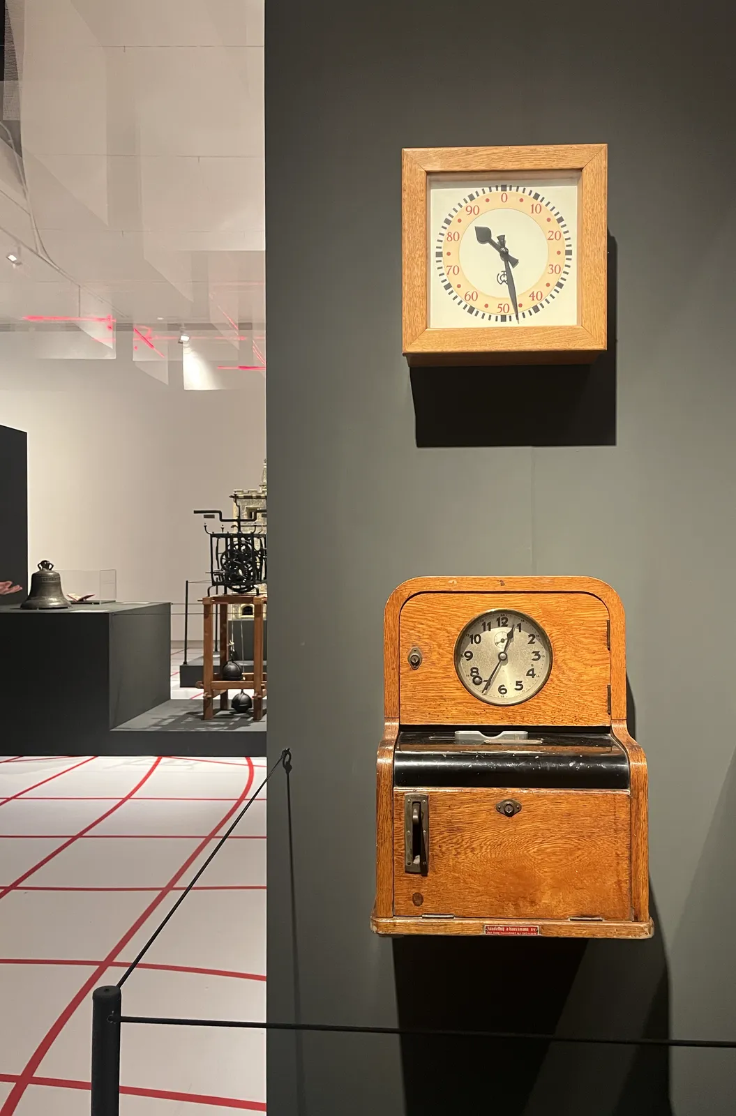 Two clocks displayed on a wall in a museum. The one on the top has the usual 60 minutes and twelve hour marks on the outside of the face, but it also has decimal indicators on the inside, which go from 0 to 99. This clock points at 28 minutes past 10 in usual time, and at approximately 47 minutes past 80 in decimal time. The other clock is an old wooden time clock, like you would see in factories. It points at 34 minutes past one, time to check out.