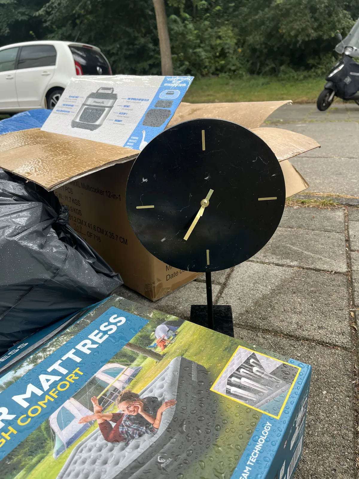 Garbage and rubbish on the street. In this pile stands a clock with skewed hands. It points at approximately 35 minutes past one.