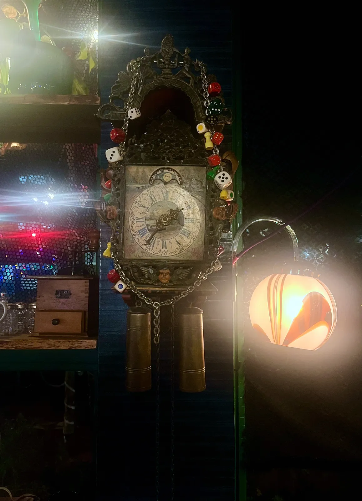 A chain with dice in all kinds of colours is hanging across this old, wooden, mechanical wall clock. Next to it there’s a seventies style glass lamp, shining brightly. The clock points at 36 minutes past one.