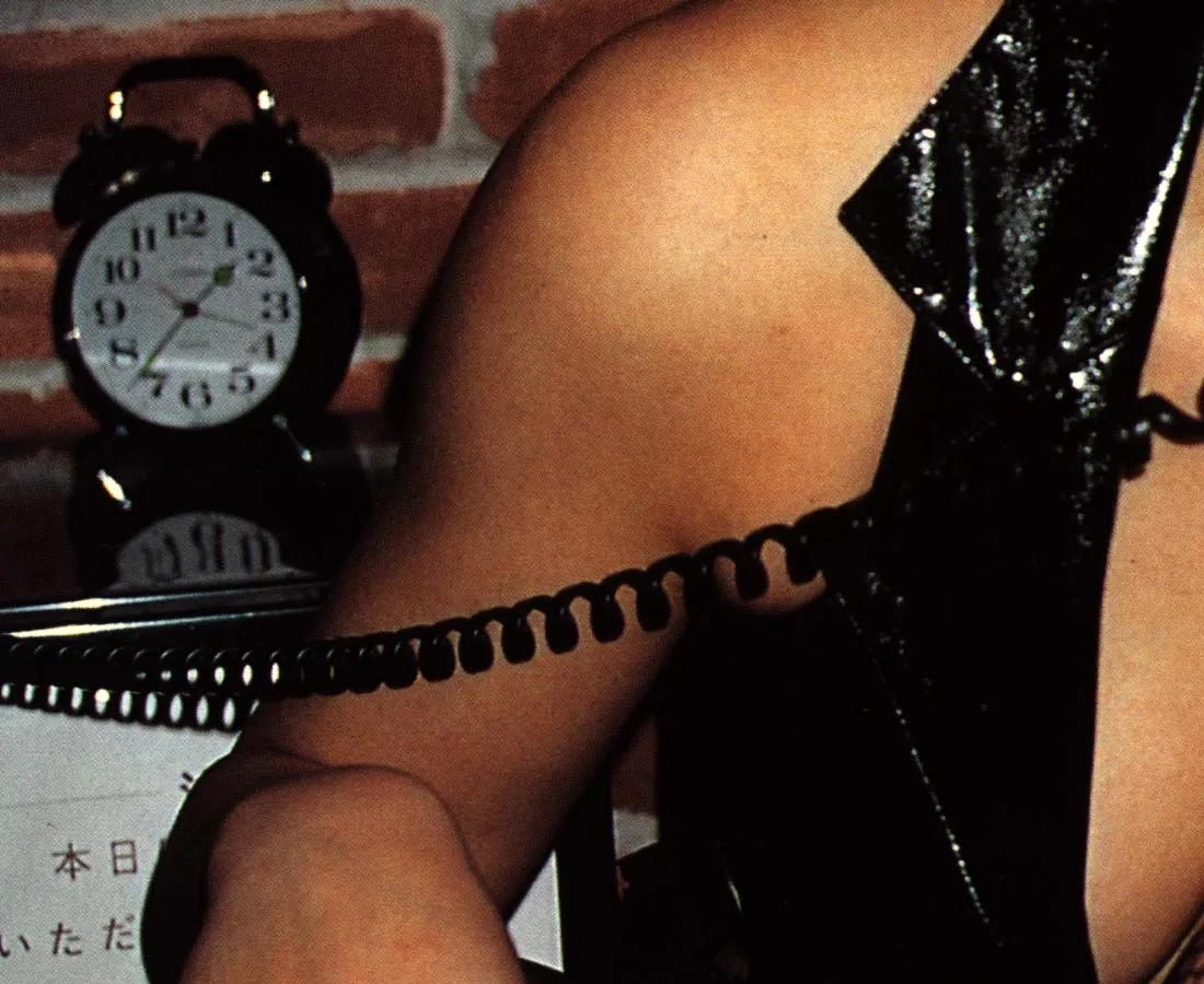 An old alarm clock behind a person in short sleeves points at thirty seven minutes past one. 