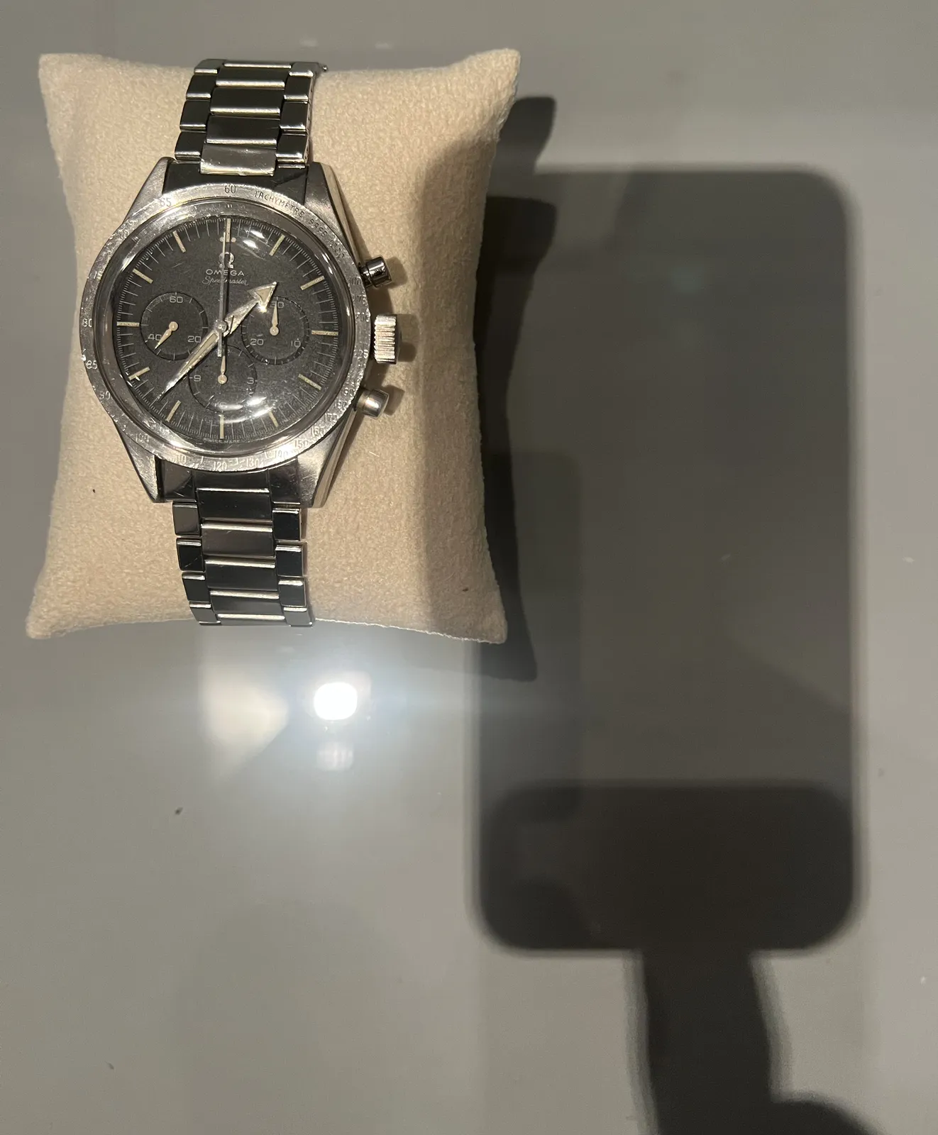 There’s a big wrist watch, attached to a little pillow in this glass showcase. The picture is taken with a phone, which is placed on the glass top, and the photographer is touching the phone with one finger. The shadow of this phone and finger take up the complete right side of the picture. On the left is this big metal wrist watch, made by Omega. It has three knobs on the right, and three little additional dials on the its face. The main dial shows the time as 37 minutes past one.