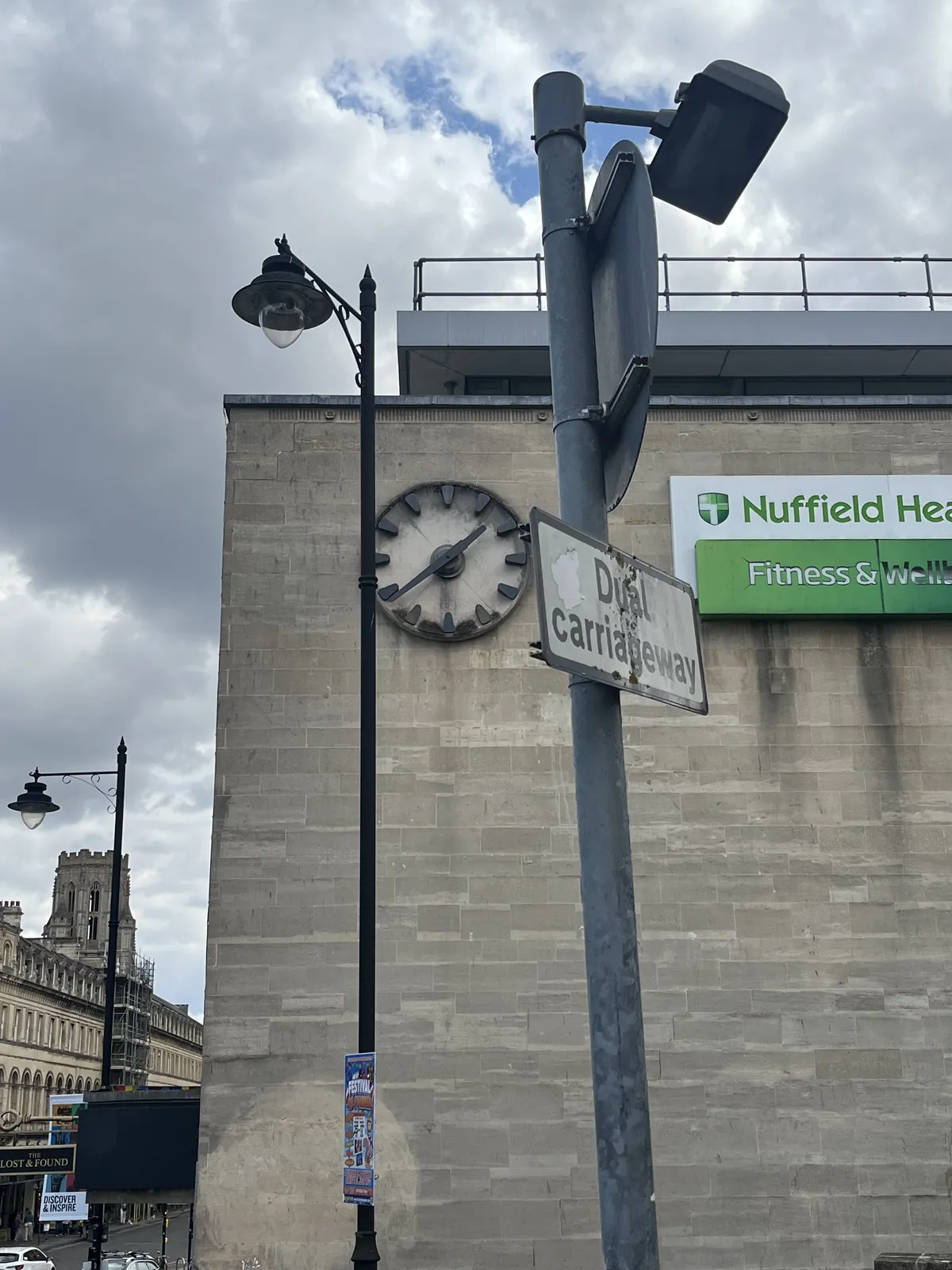 A clock built into a modern facade. There’s a pole in the foreground of the image with a sign on it that says Dual carriageway. Next to the clock there’s a new, green sign. Only the words Nuffield and Fitness are readable, the rest is out of frame. The clock points at 22 minutes to two.