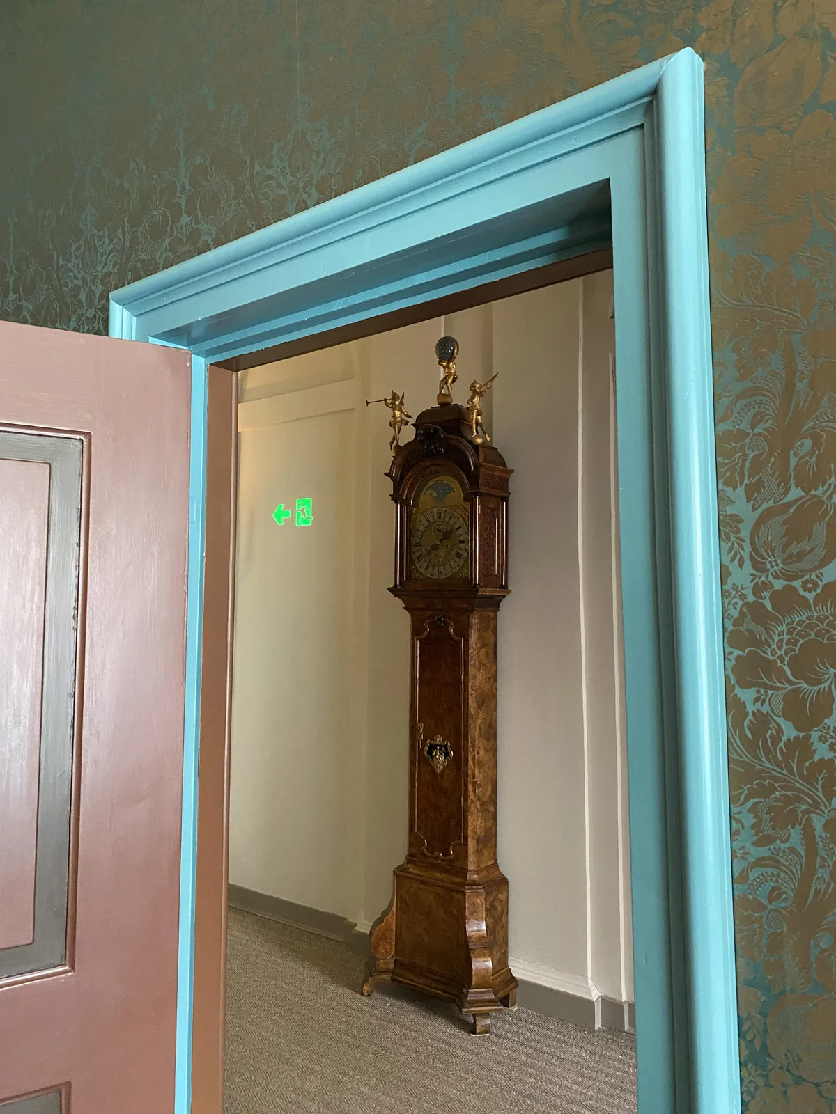 Peeking thru a door with a frame painted in a striking colour. There’s an old standing clock. It points at approximately 38 past 1.