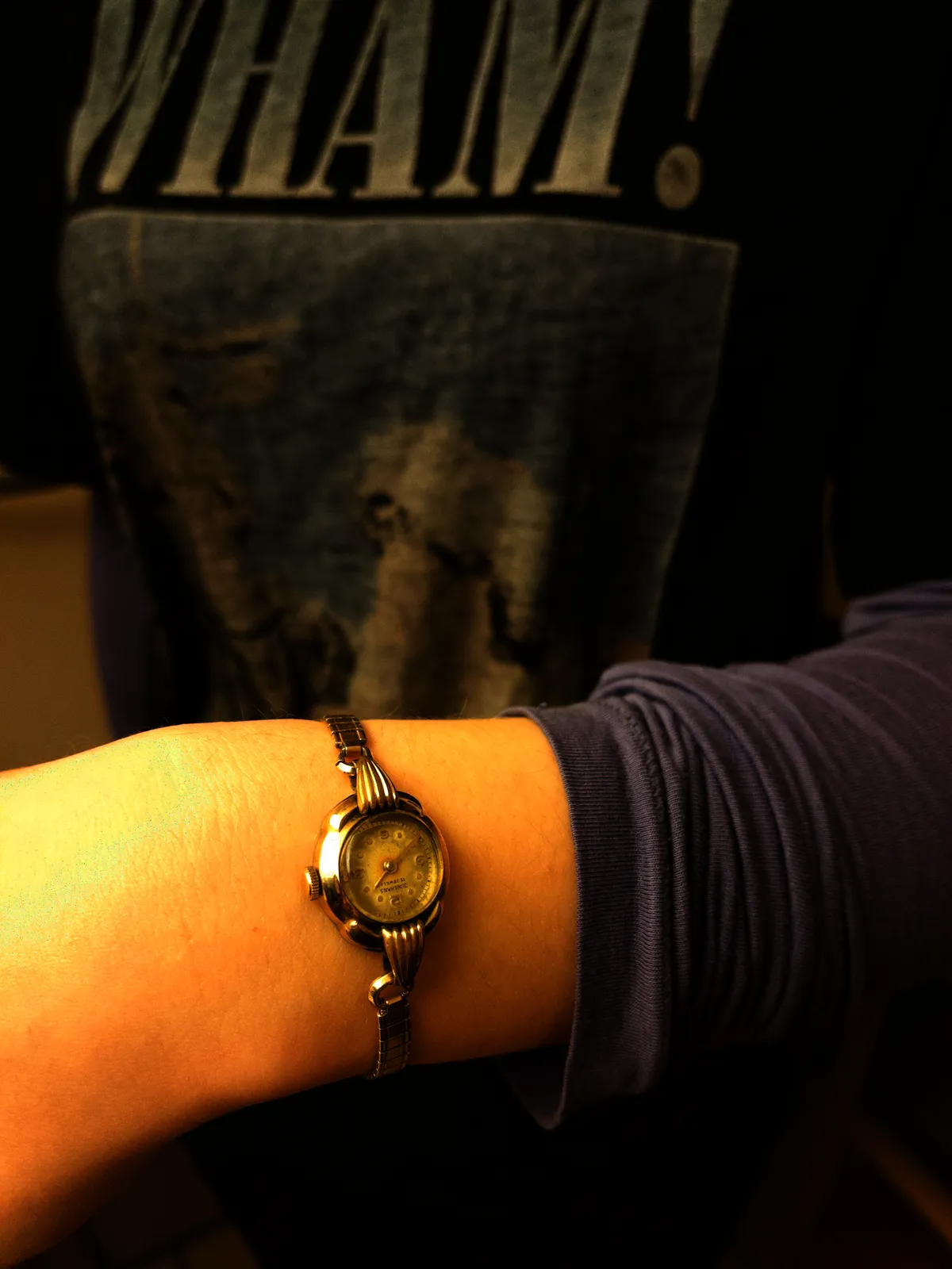 Someone is wearing a Wham t-shirt, and a small elegant watch on their wrist. It points at 39 minutes past one.