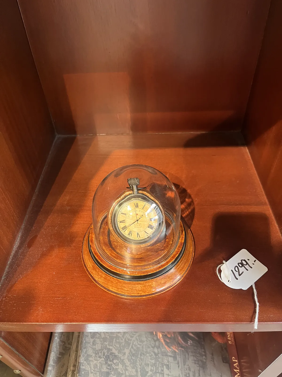 An old mechanical pocket watch in a glass dome. It is for sale for 1299 and it points at approximately 39 minutes past one