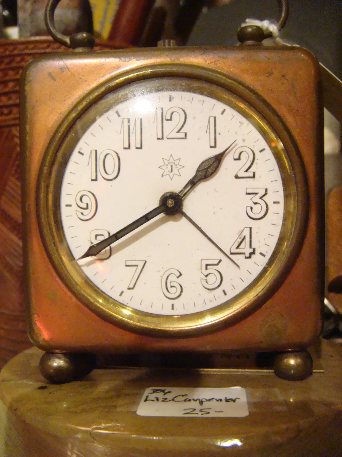 An old, square clock that seems to be made from some sort of metal. It’s for sale for 25, and it points at 20 minutes to 2.