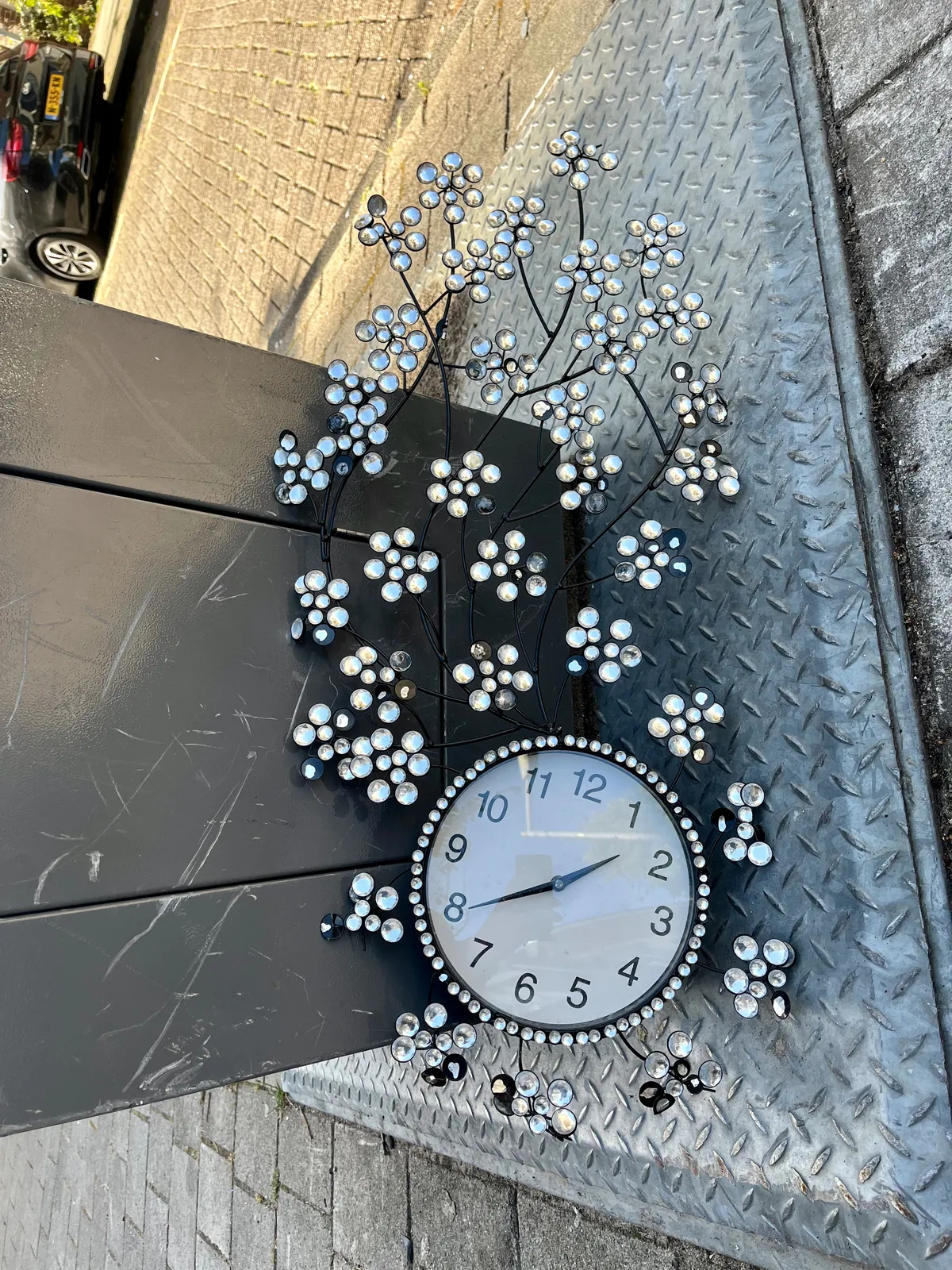 A clock, surrounded by flowery ornaments, is dumped on the street. It points at 40 past one.