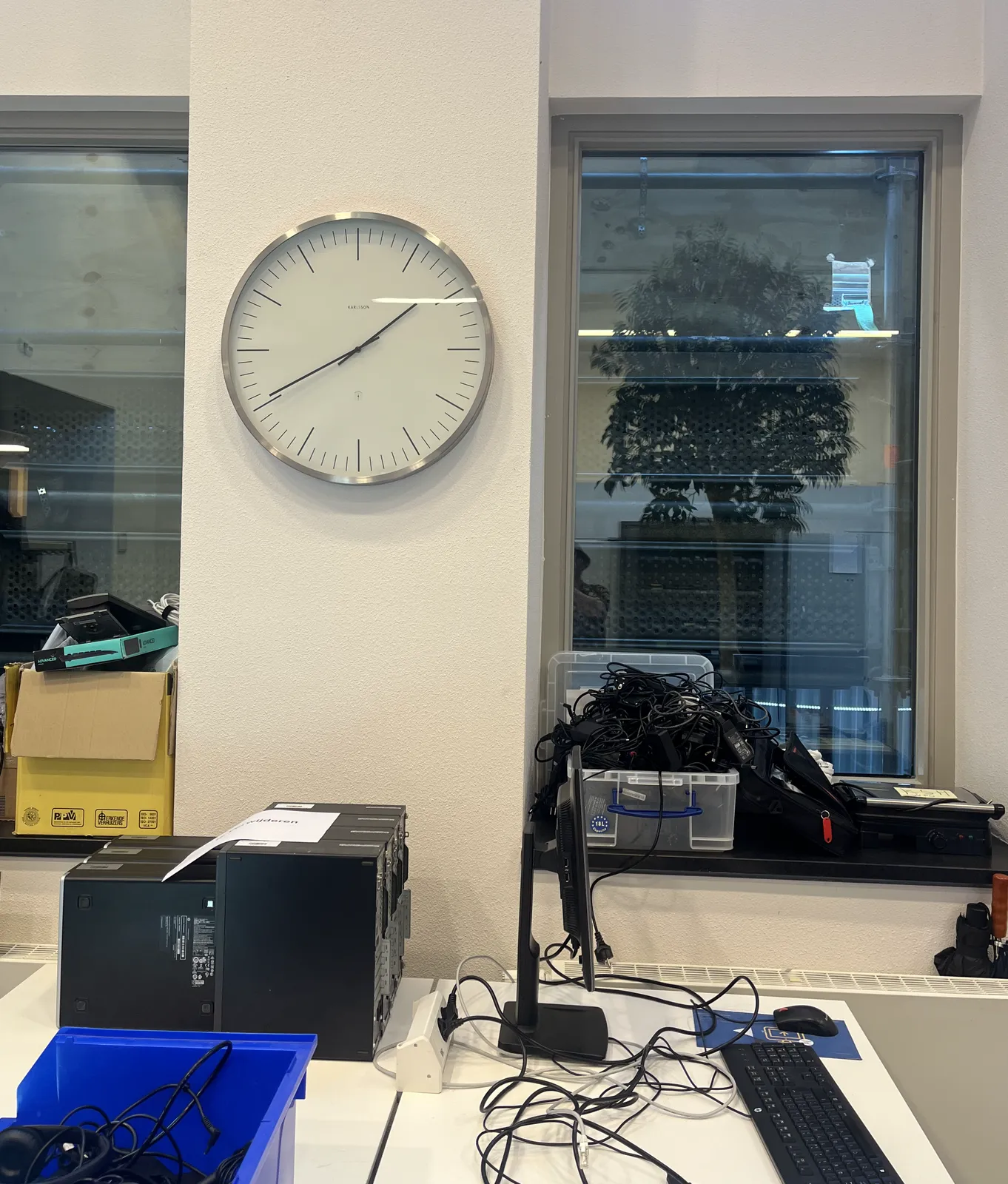 A large wall clock between two windows in an IT office. Boxes with cables, a stack of desktop computers, keyboards everywhere. It points at 20 minutes to two.