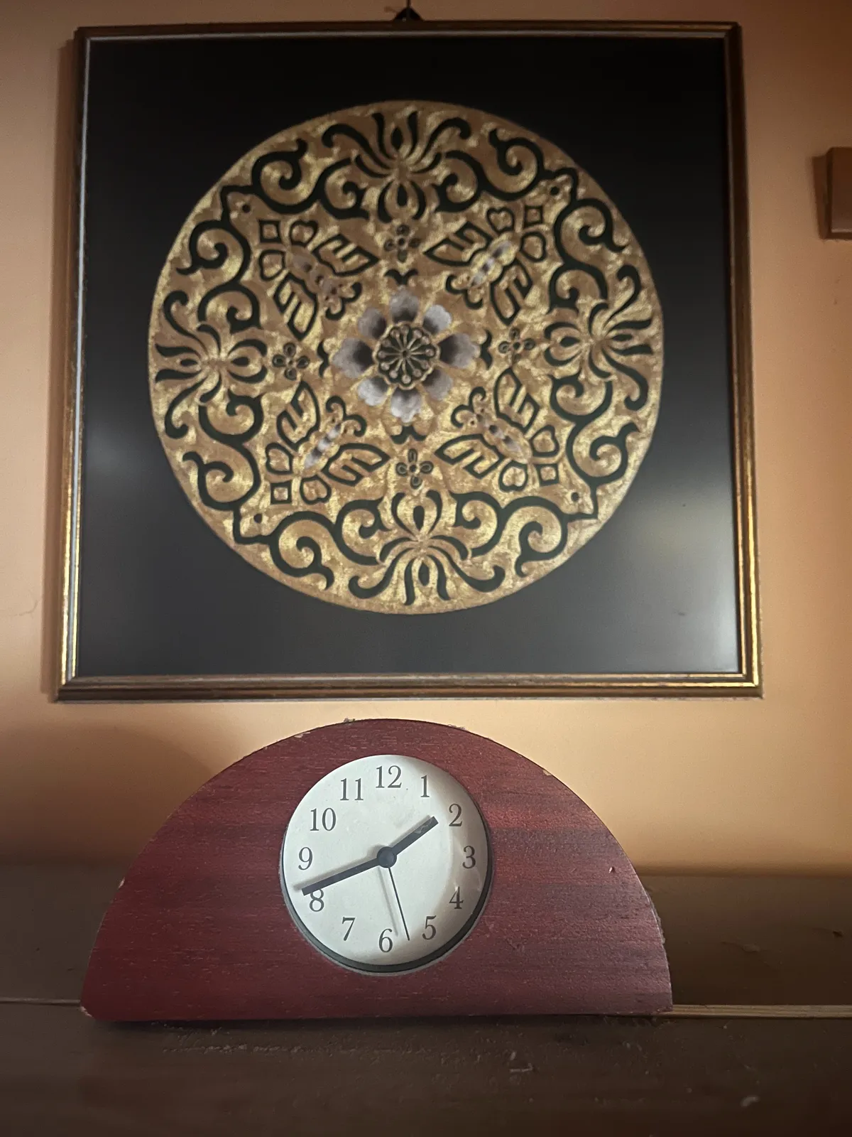 Below a gold on black circular, flowery illustration, there’s a wooden clock, in the shape of a semi circle. This clock points at 19 minutes to 2