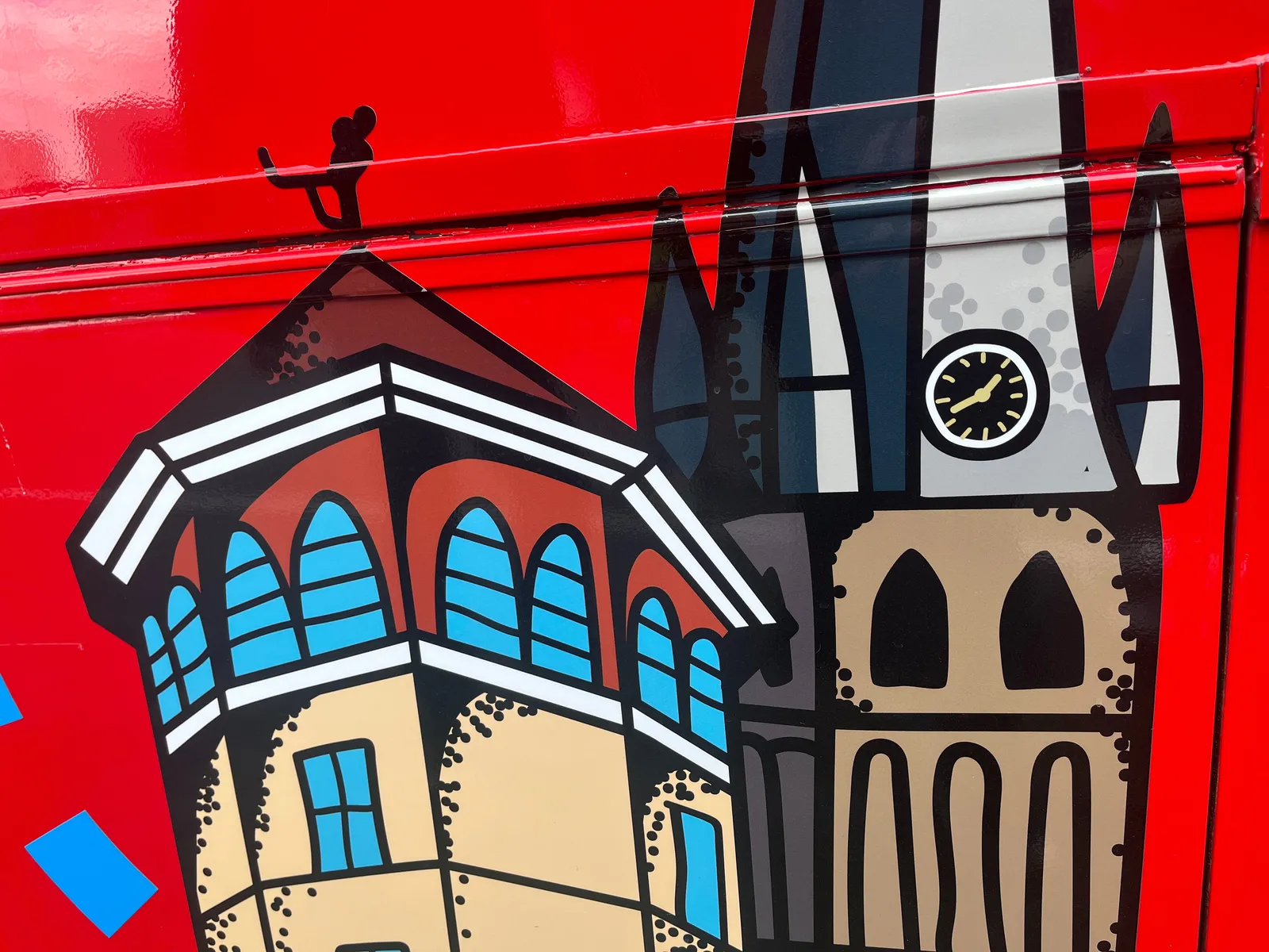 Looking at a detail of a drawing of a city, stickered onto a red car. On this drawing there are two buildings, one of them is a pointy church tower with a clock on it. It points at approximately 18 minutes to two  