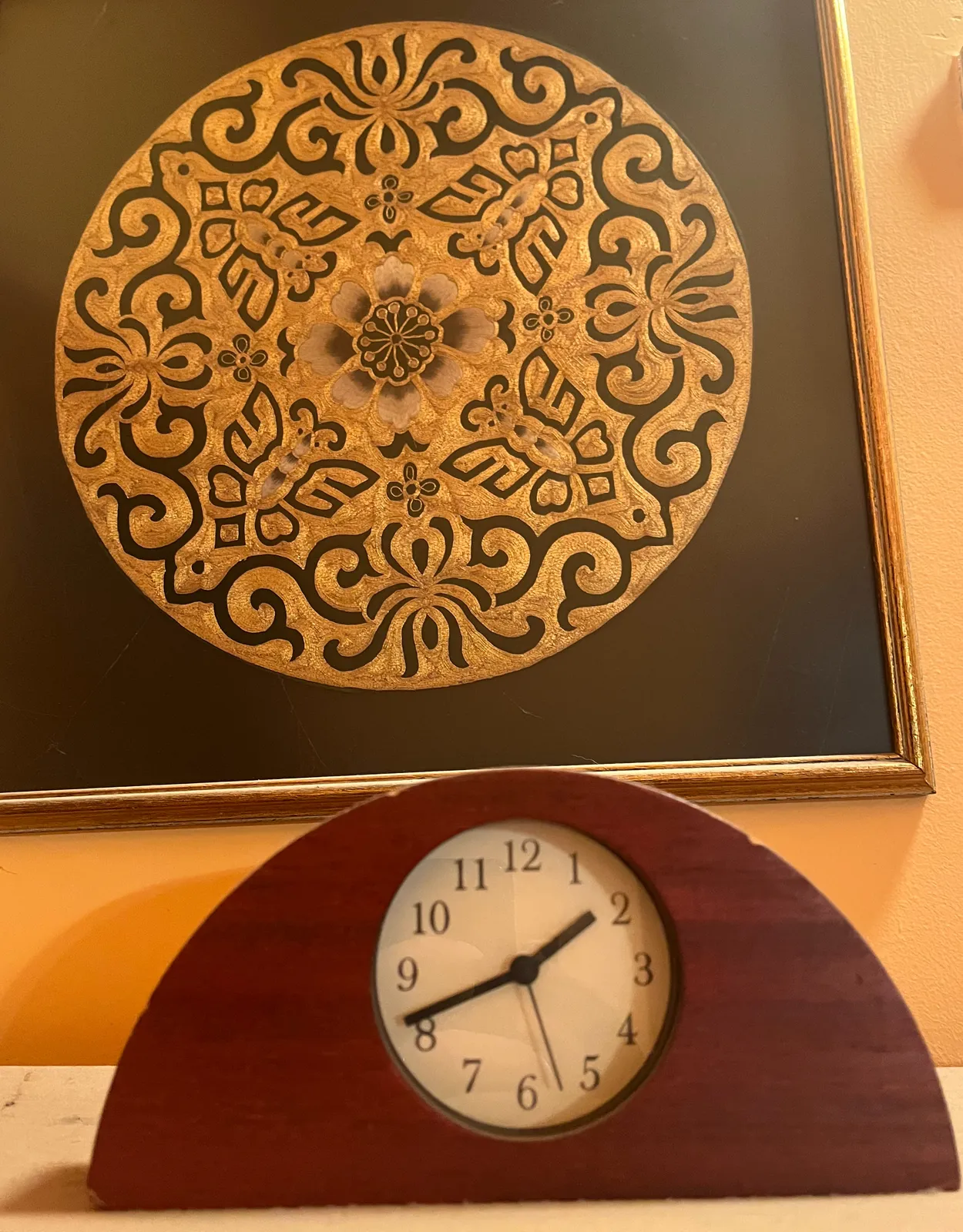 A frame with a circular, gold embroidery in it. Below it is a wooden clock. And on it it is 18 minutes to two.