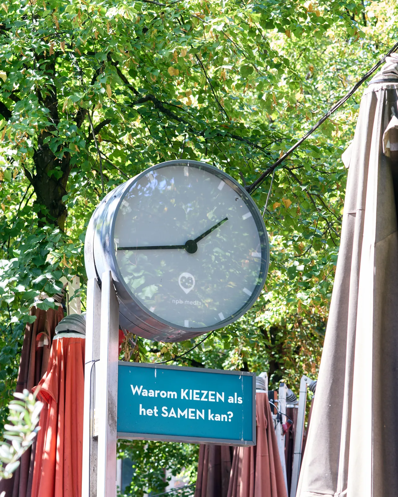 A gray clock on a pole with green trees behind it. The clock points at 16 minutes to 2. Below the clock there’s a sign that asks the question, in Dutch: Why choose when you can do it together?