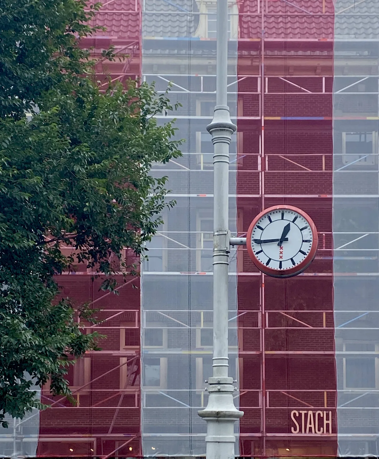 A building in scaffolding. There’s an enormous sheet draped over it, with wide, vertical white and red stripes. In front of this building there’s a tree, and a clock attached to a pole. This clock points at 16 minutes to 2.