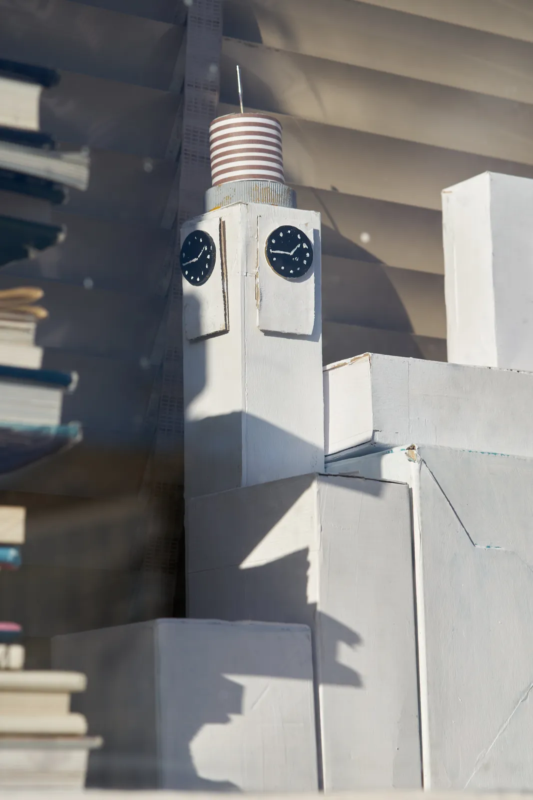 Next to a pile of books there’s a scale model of a white, modernist building. It has a tower and on that tower there’s a black and white clock. It points at a quarter to two.
