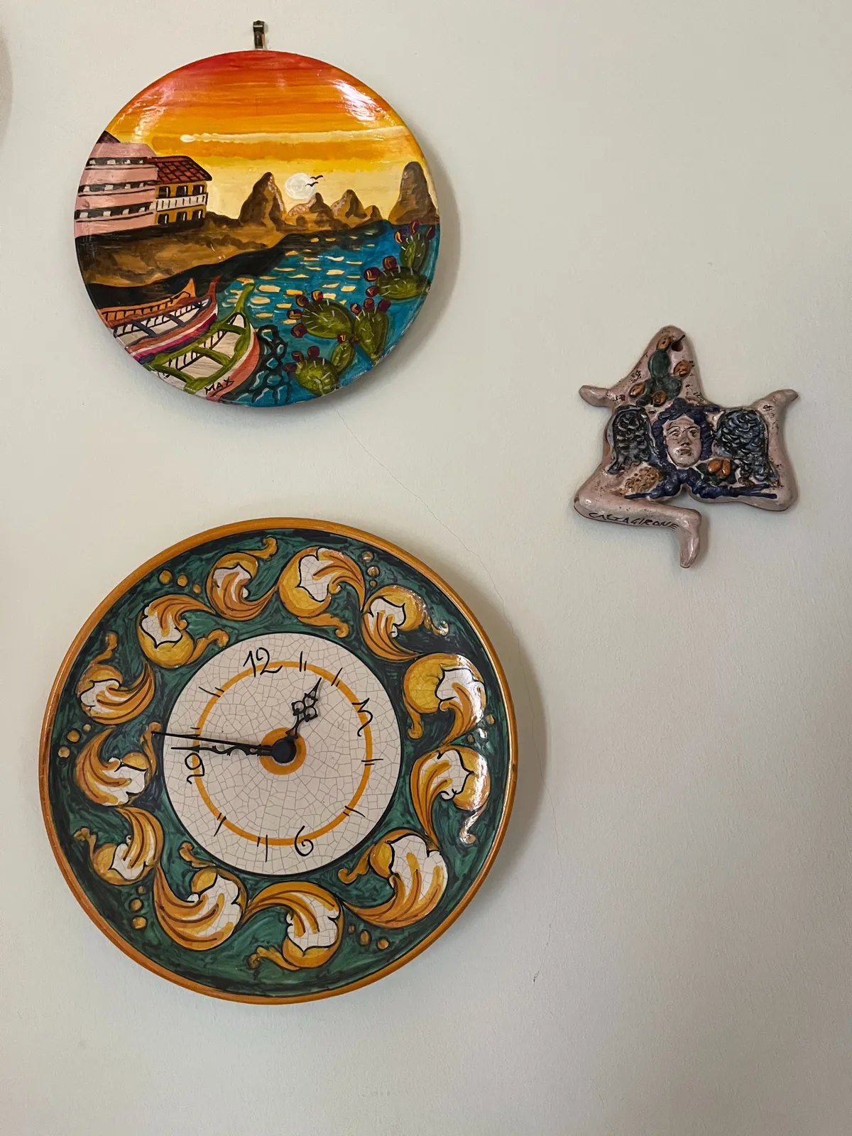A ceramic version of the symbol of Sicily, a three legged monster, if hanging on a wall, next to two colourful ceramic plates. The one at the top has an image of some houses, some boats and a sun setting behind some mountains on it. The other plate is ornamented with pepper like shapes. This plate is also a clock which points at 14 minutes to 2.