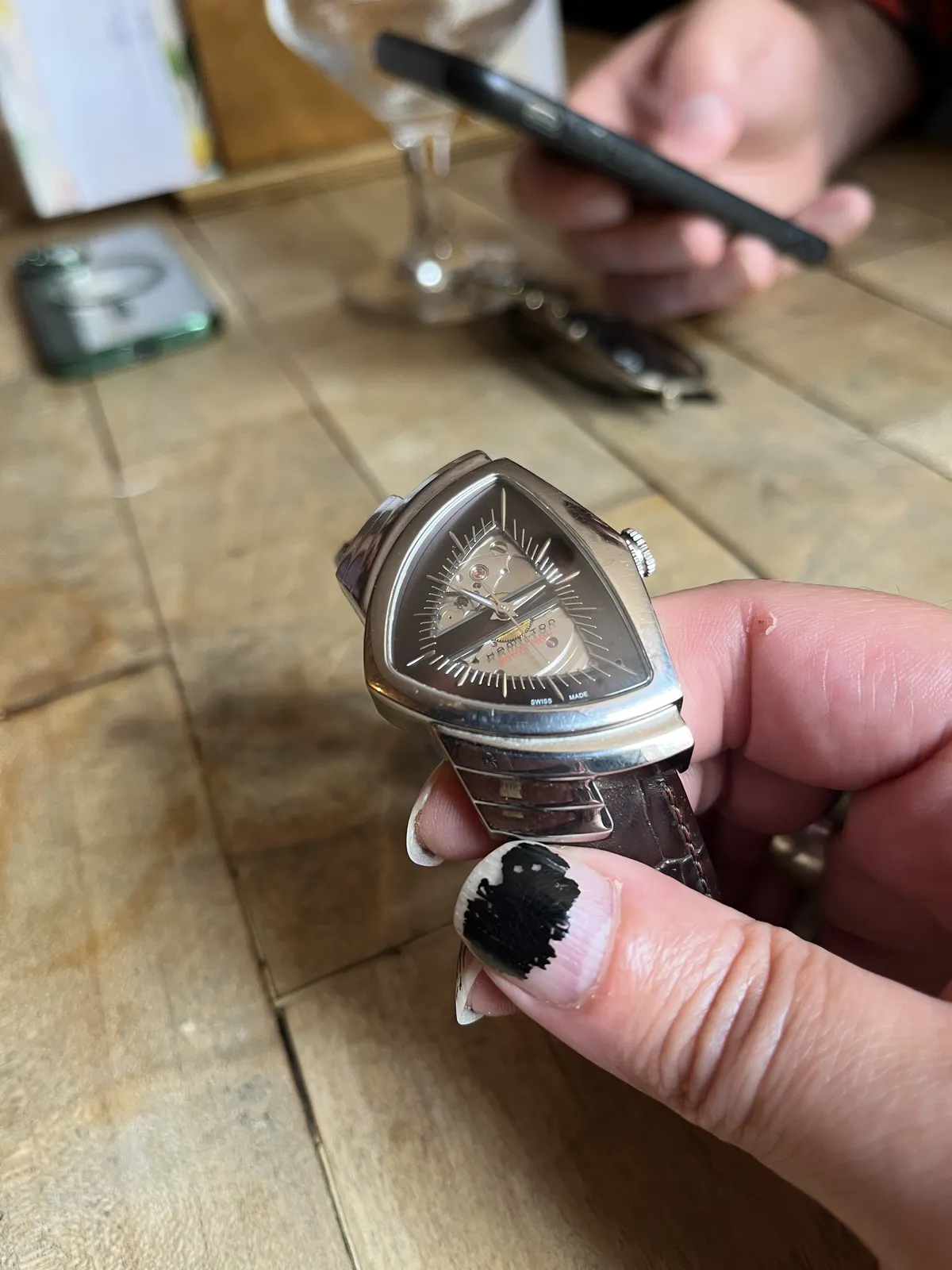 The background is blurry, but you can see someone’s hand holding a phone, and another phone lying on the table. In the foreground, someone with long nails, with severly worn nailpolish, holds a wrist watch in their hands. It has a triangular shape, very well designed. Since it’s triangular it is not straightforward to read the time, but once you get it you can see that it is ten minutes to two. 