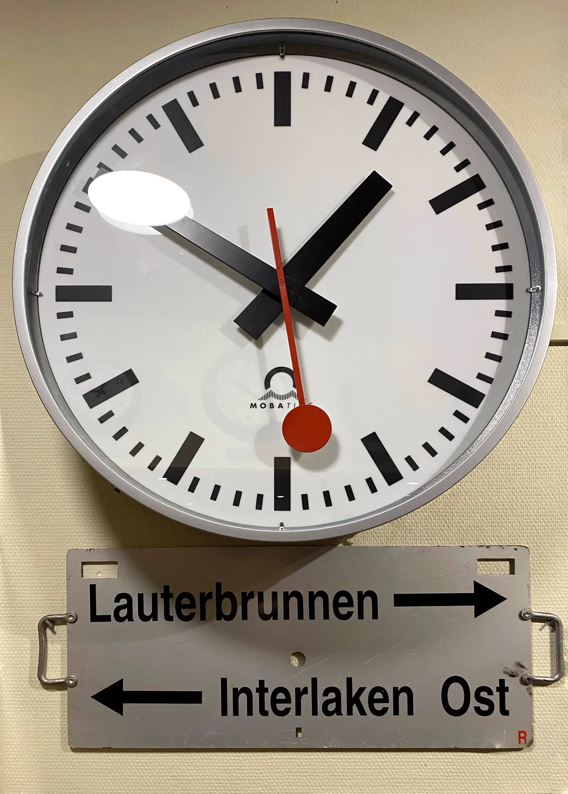 A sign that says that Lauterbrunnen is to the right, and Interlaken Ost is to the left. Above this sign there’s a minimalist Swiss clock, with a red second hand with a circle at its tip. This clock points at 10 minutes to 2.