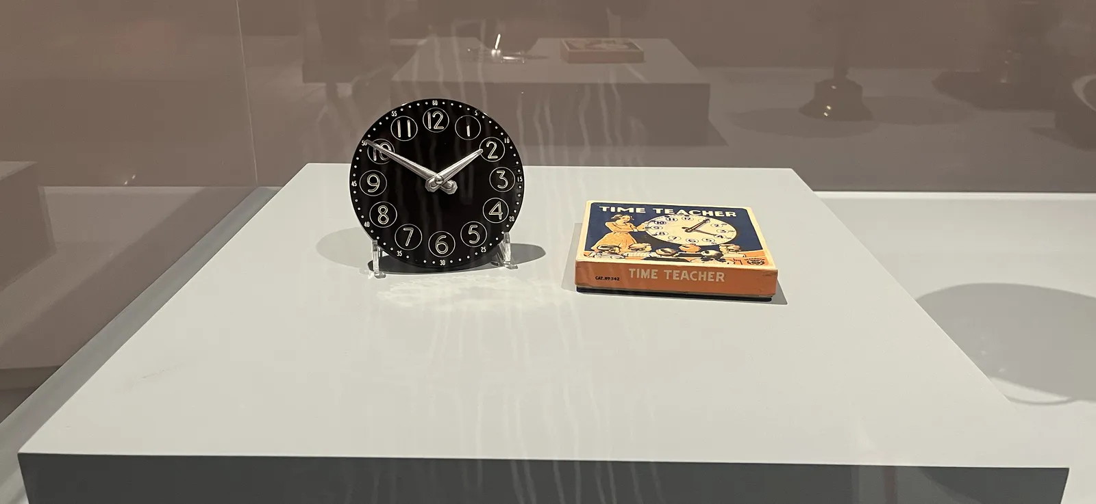 In a glass showcase there’s this black and white clock, its hands point at 10 minutes to two. Next to this clock lies the box of this clock. The words Time Teacher are printed on it, above an image of this same clock, but this one points at 20 minutes past one instead.