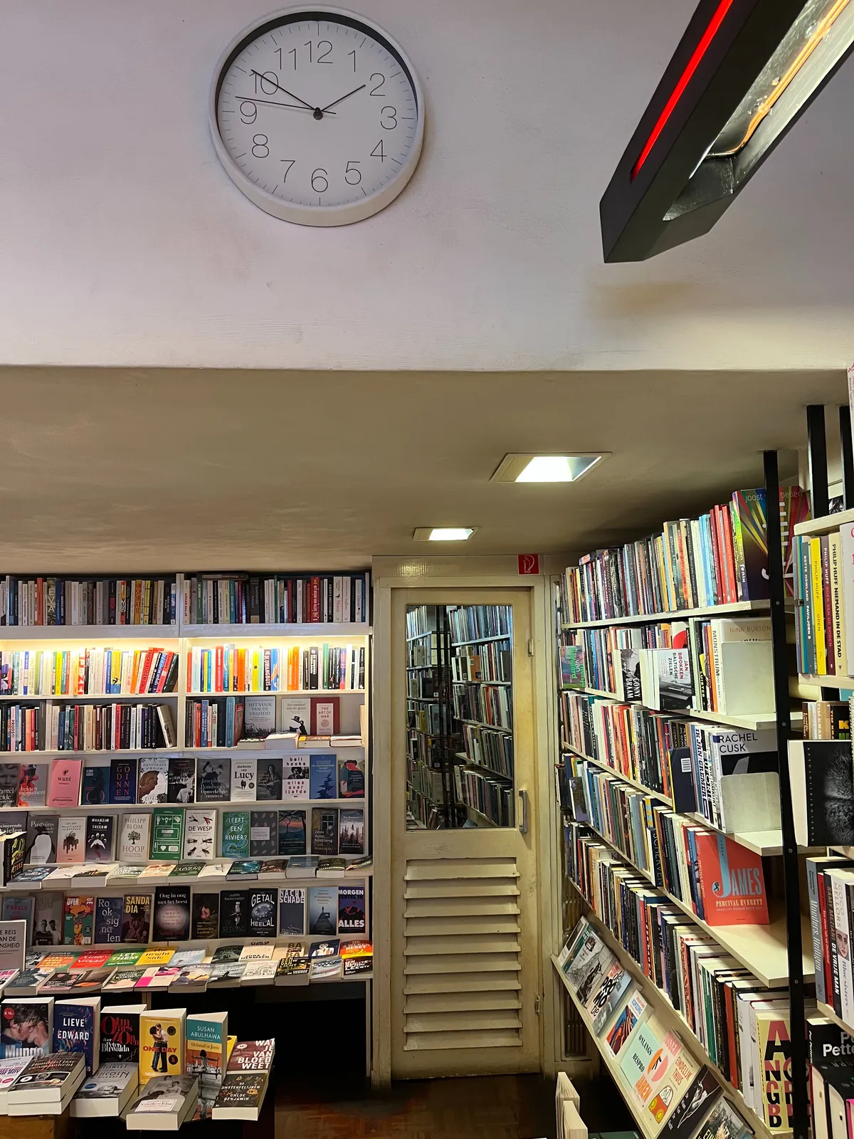 A simple modern wall clock hangs on a wall in a book store. There are lots and lots of books. The clock points at 10 minutes to 2.