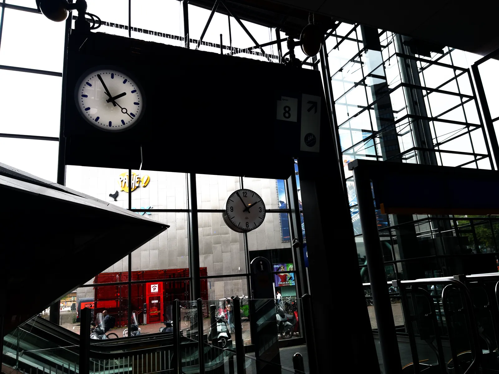 Looking at a digital train display, and at the glass facade of a modern Dutch train station. In the facade there’s a clock, and there’s a clock attached to the display as well. They both point at 5 minutes to two.