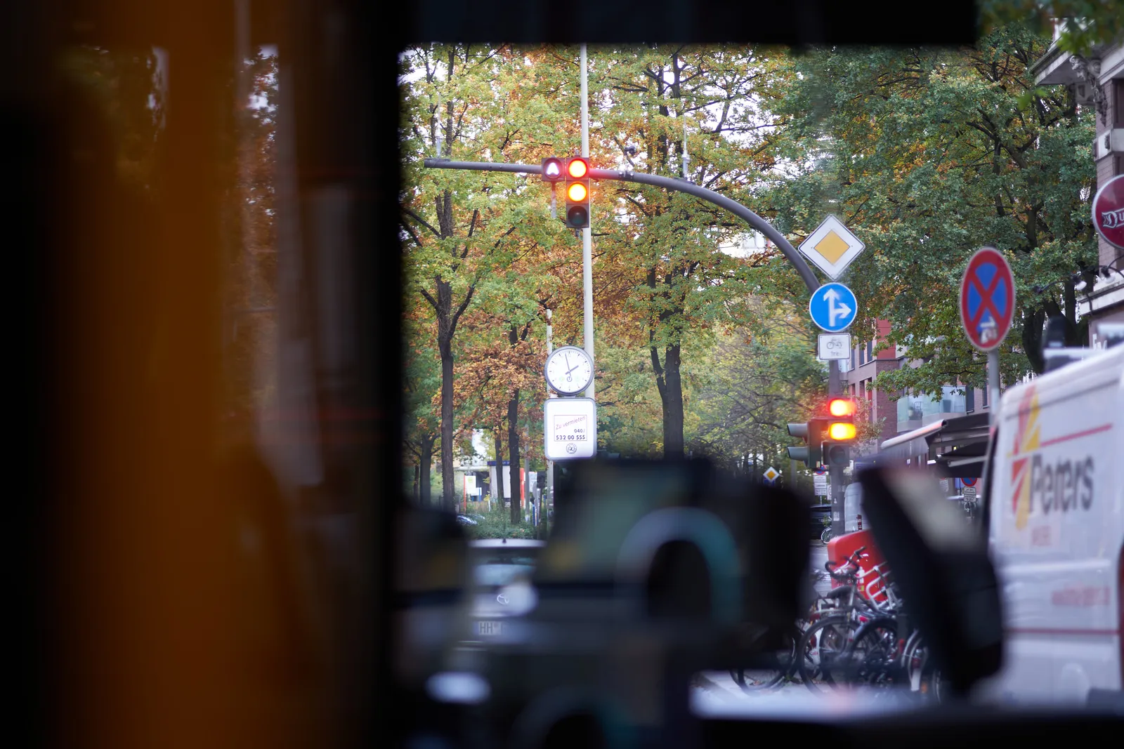 Looking through the front window of a bus. There are some trees, cars, traffic lights, and a clock. The clock points at 2 minutes to 2.