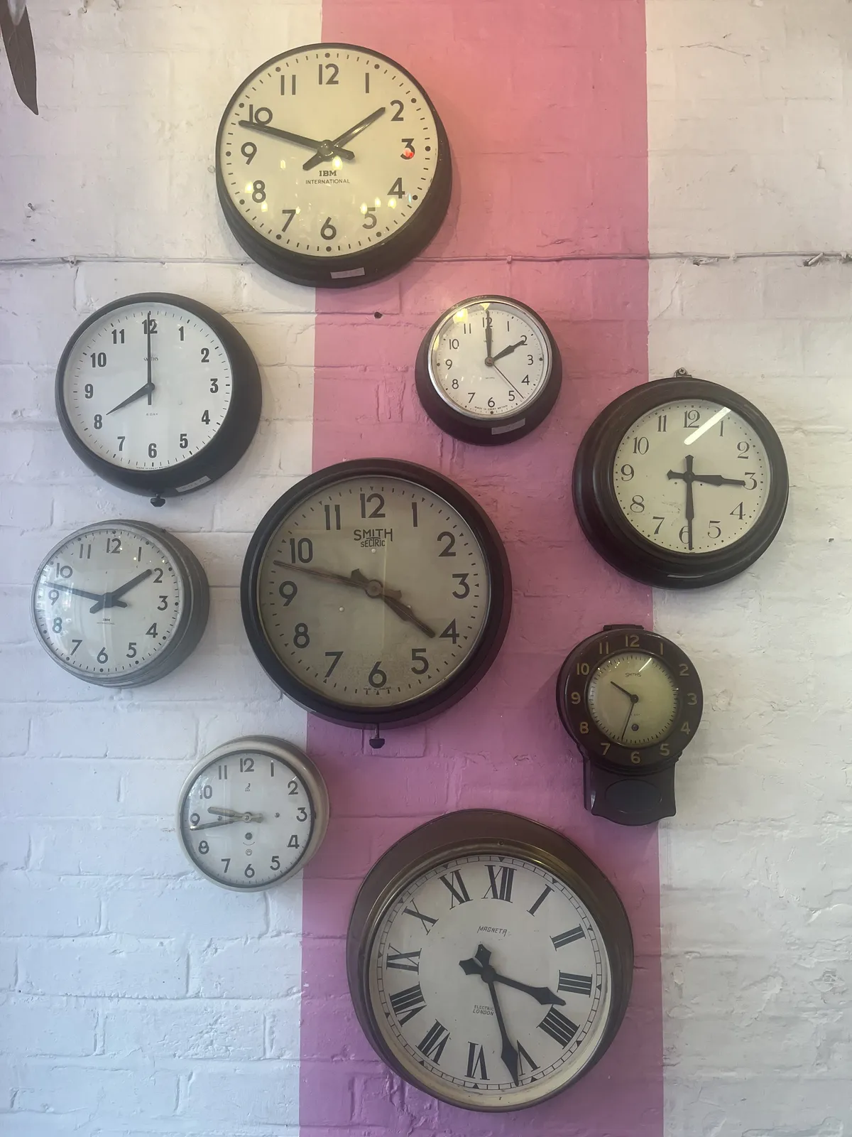 There are nine wall clocks on this brick wall. The wall is painted white, with a pink band in the middle of the image. The clocks are carefully hung onto the wall in a slanted square shape. One of the clocks points at 13 minutes to two. There’s one that points at 12 minutes to two. There’s another one that points at exactly two o’clock. Another clock points at 26 minutes past three, and there’s one that points at 29 minutes past three. Another one points at 12 minutes to five. There’s one that points at one minute to eight. Another clock points at 17 minutes to ten. And the last one points at 33 minutes past 10.