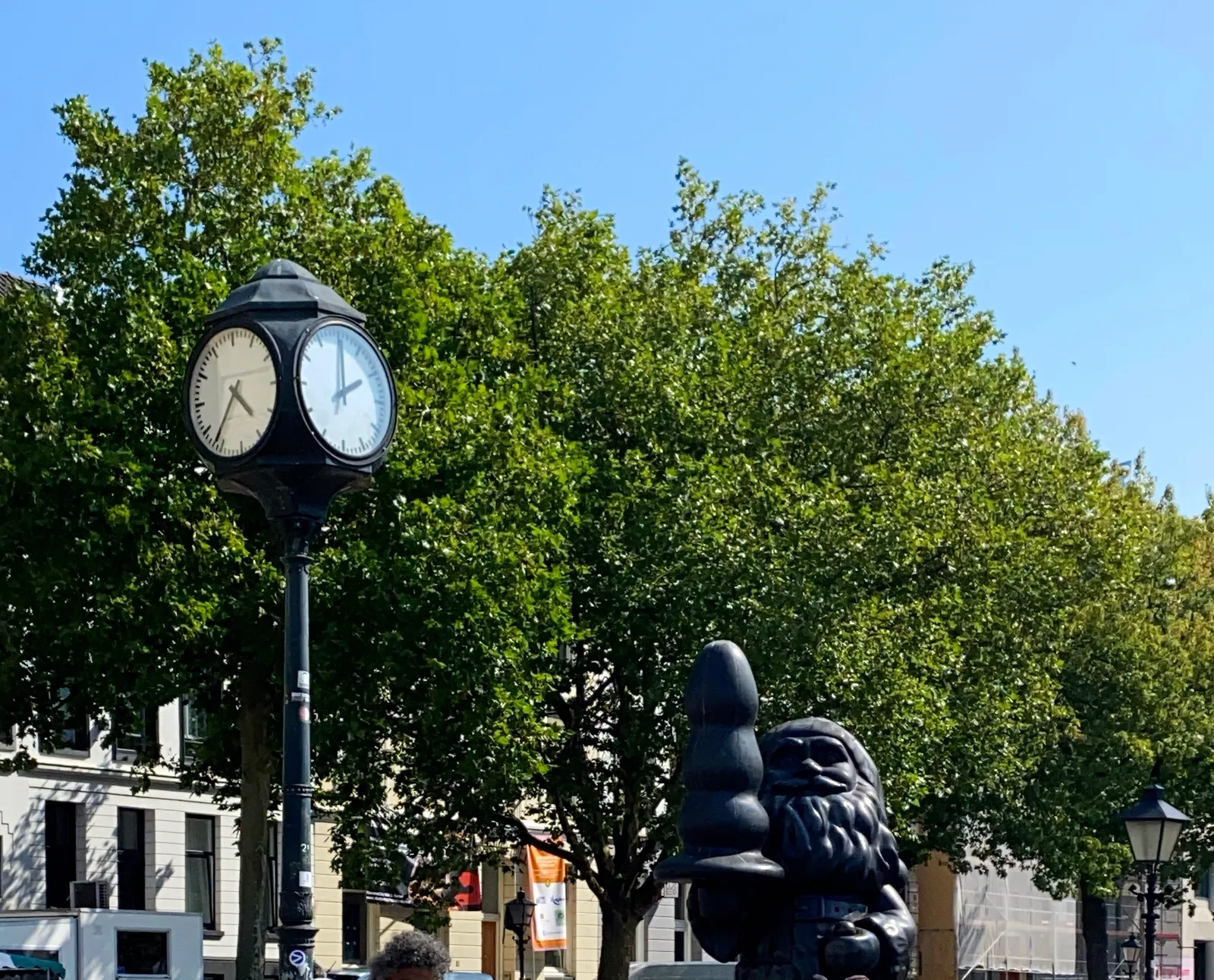 A sunny day, with trees with leaves. The famous statue of a gnome with a large butt plug in his hand is in this picture. There’s a pole which has clocks on four sides. The clocks on two sides can be seen. They point at different times. The one on the right points at 2 o’clock, the one on the left at 36 minutes past 4.
