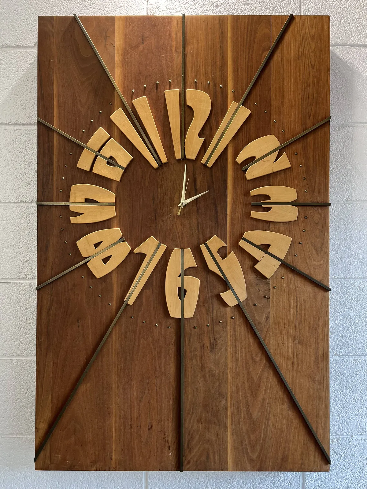 A wonderfully designed, wooden clock. It’s rectangular in shape. The hour indicators are formed by long strokes that extend to the sides of the frame. The numbers are made from art nouveau style type. It points at one minute past 2.