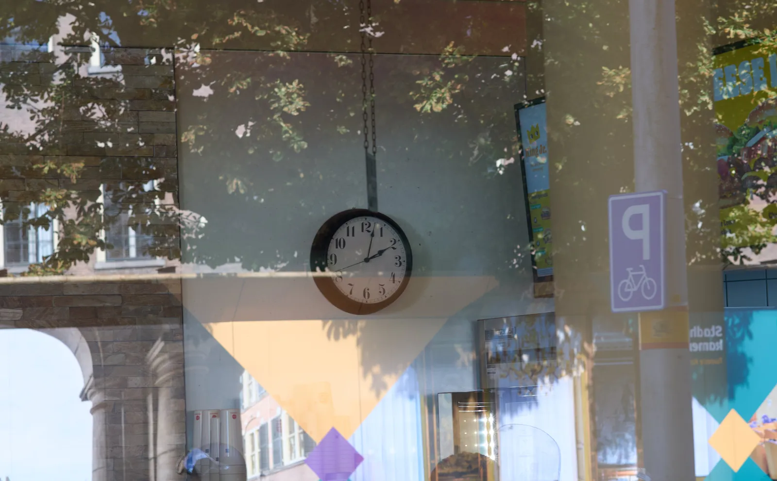 Peeking through a shopping window. In the reflection a tree can be seen, and a neoclassical arc and a column. Inside the shop there’s a clock on the wall that points at 2 minutes past 2.