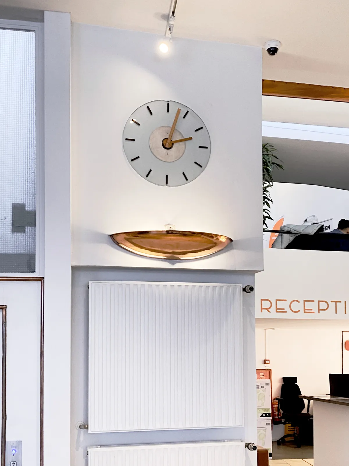 A designed clock built into a wall in, what seems to be, the reception hall of a hotel. The clock points at 3 minutes past 2. 