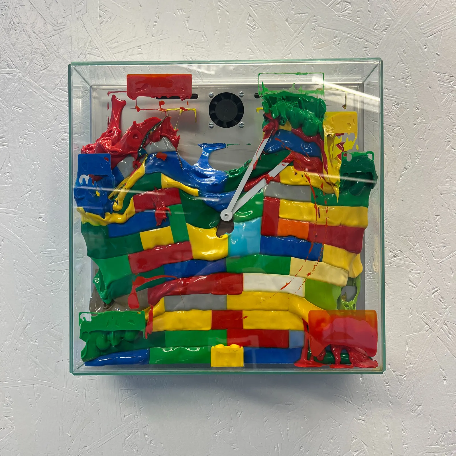 A clock in a transparent plastic box. It’s very colourful. Is seems to be made from large, melted lego/duplo blocks. The hands are stuck in the molten blocks, and the point at 4 minutes past 2 forever.