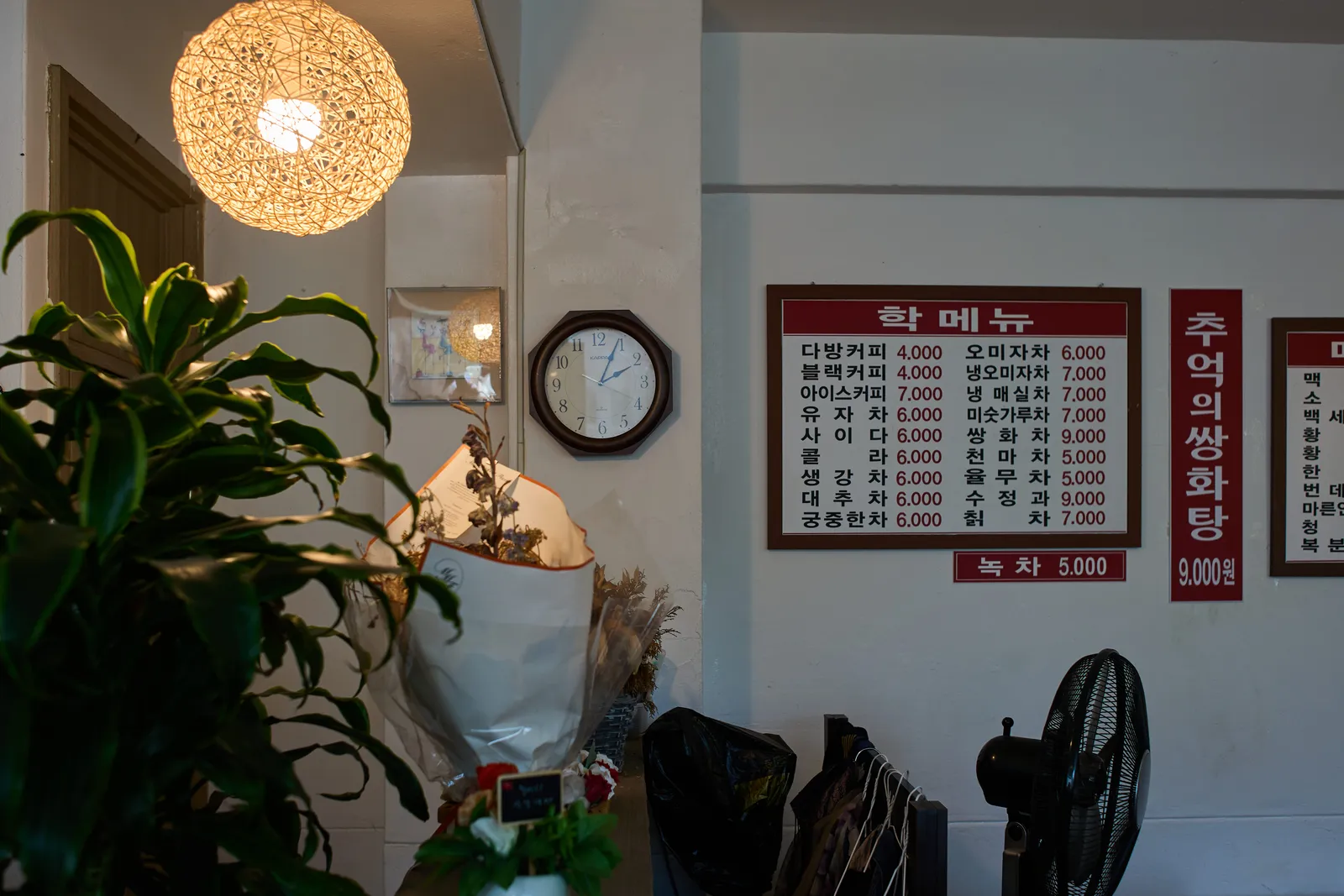 There are plants on the left, and a clock in the middle of the picture, hanging on the wall. It seems to be a Korean restaurant, by the looks of the large menu, pinned to the wall, next to the clock. The clock points at 4 minutes past 2.
