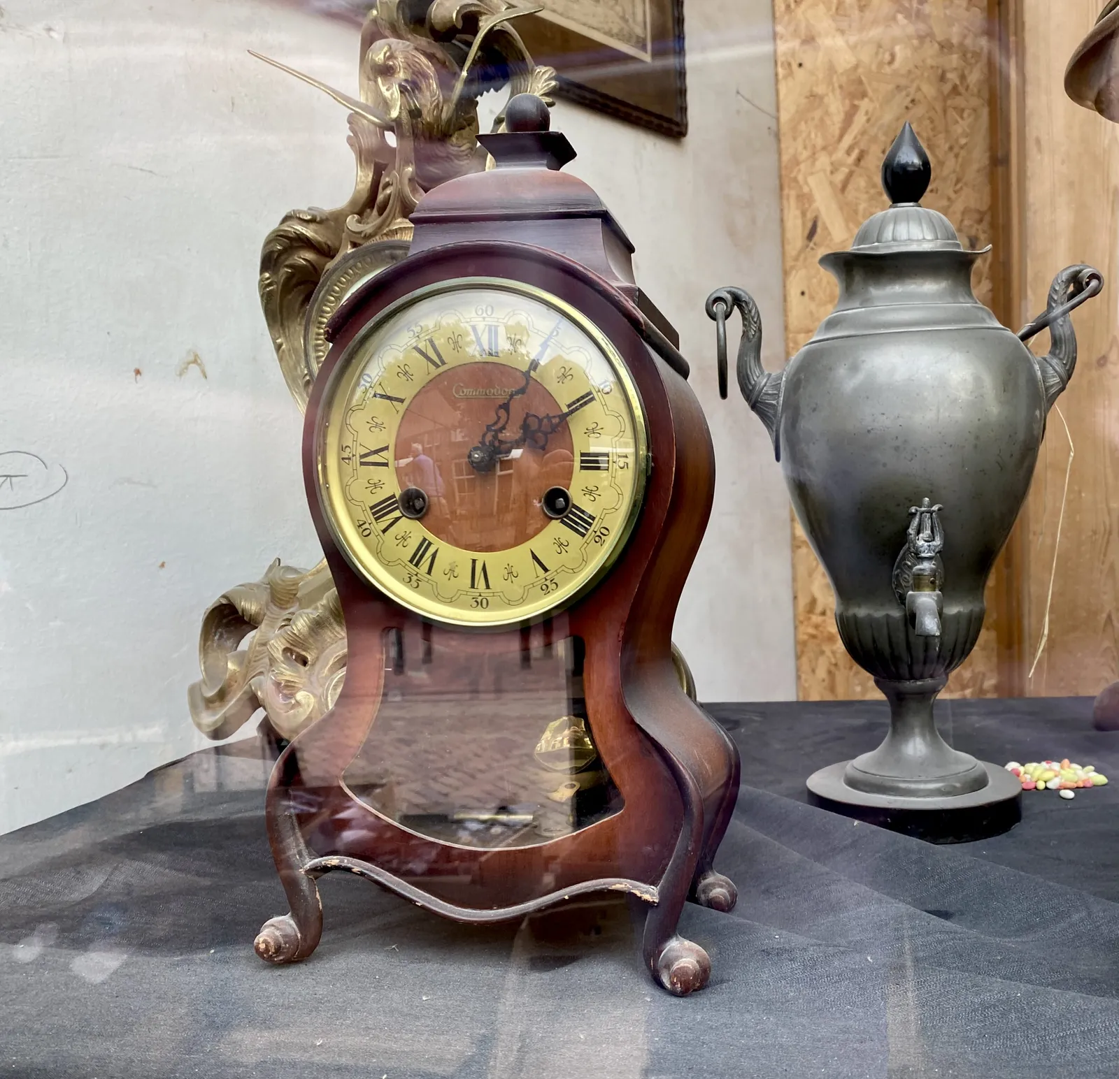 A metal, ornamented vase, with a little tap. Next to it is an old, elegant desk clock that points at 5 past 2.
