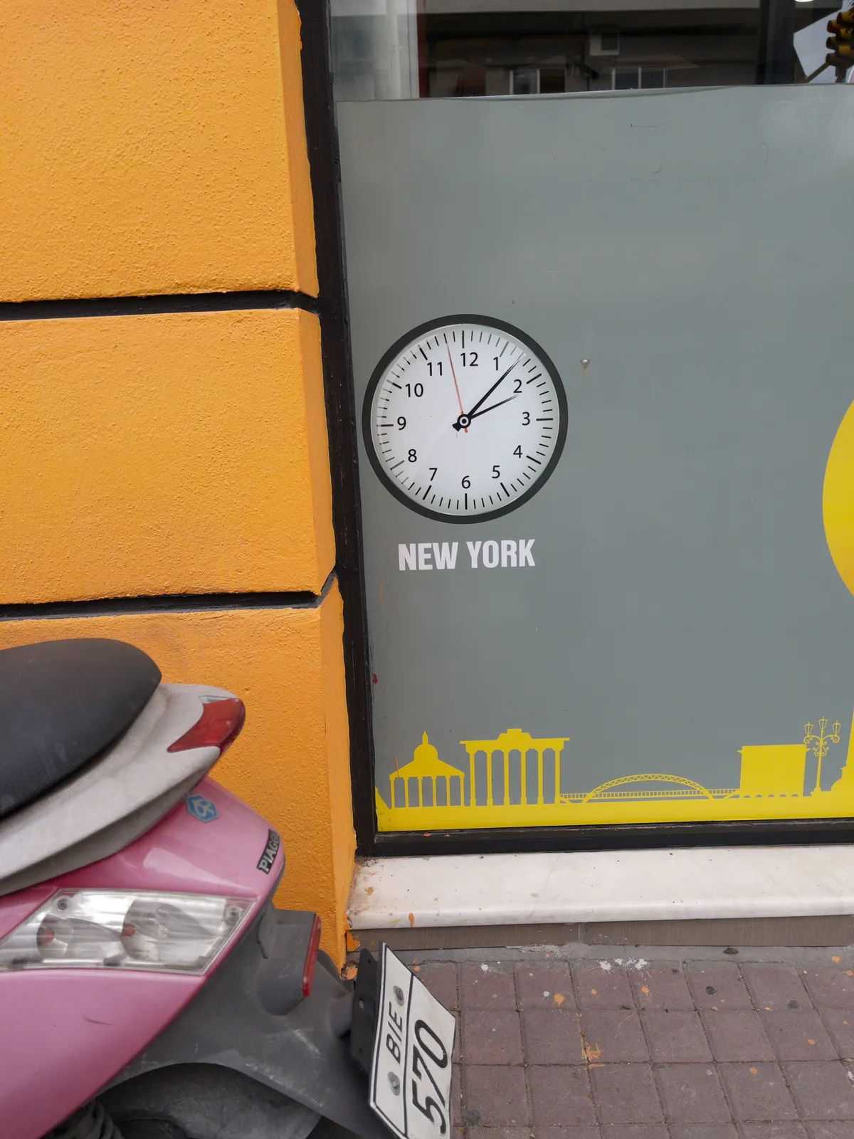A pink moped is parked in front of an organge-yellow wall. Next to the wall is a large window with a clock printed on it. Below the clock it says New York. The clock points at 7 past 2