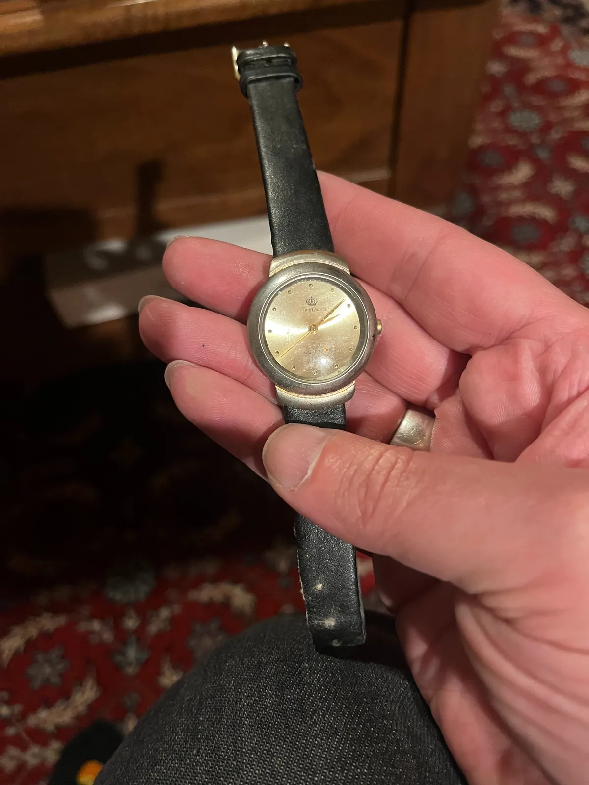 Someone is holding an old watch in their hand. In the background, blurry, an old Persian rug, and a dark wooden chest. The watch points at 8 minutes past 2