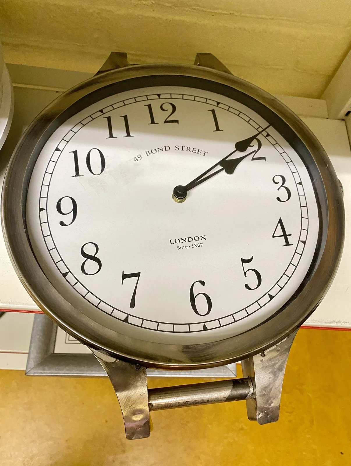 A large wall clock (laid down on a shelf) in the shape of a wrist watch (without bands). 49 Bond Street London Since 1867 on its face. Numerals with a serif, and classic hands that point at 9 minutes past 2.