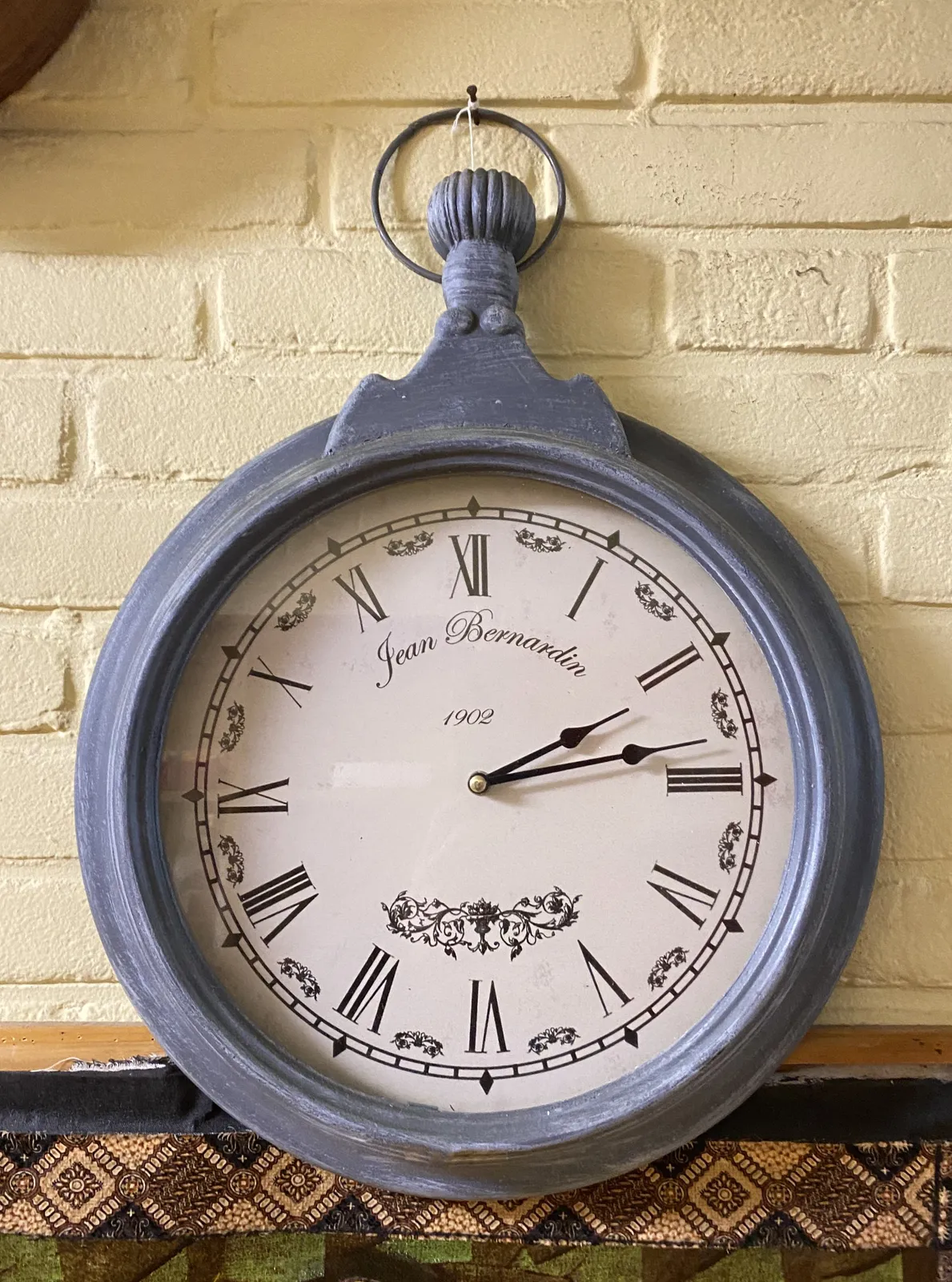 A wall clock shaped like an old fashioned pocket watch. Roman numerals, some floral illustrations, and the words Jean Bernardin 1902 printed on its face. The classic hands point at 13 minutes past 2.